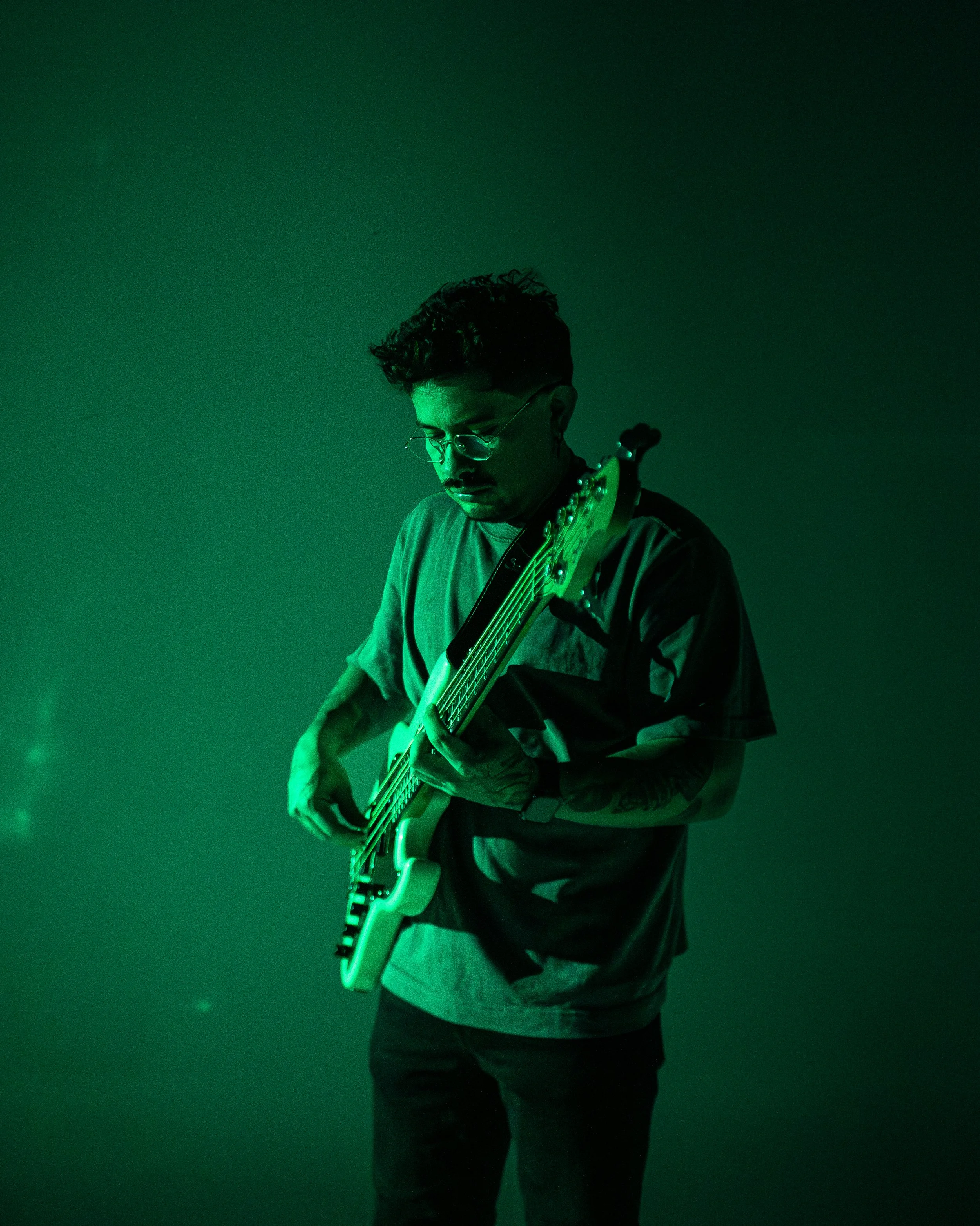 A person with curly hair, glasses, and tattoos wearing a dark sweatshirt, standing against a dark green background, holding an electric guitar.