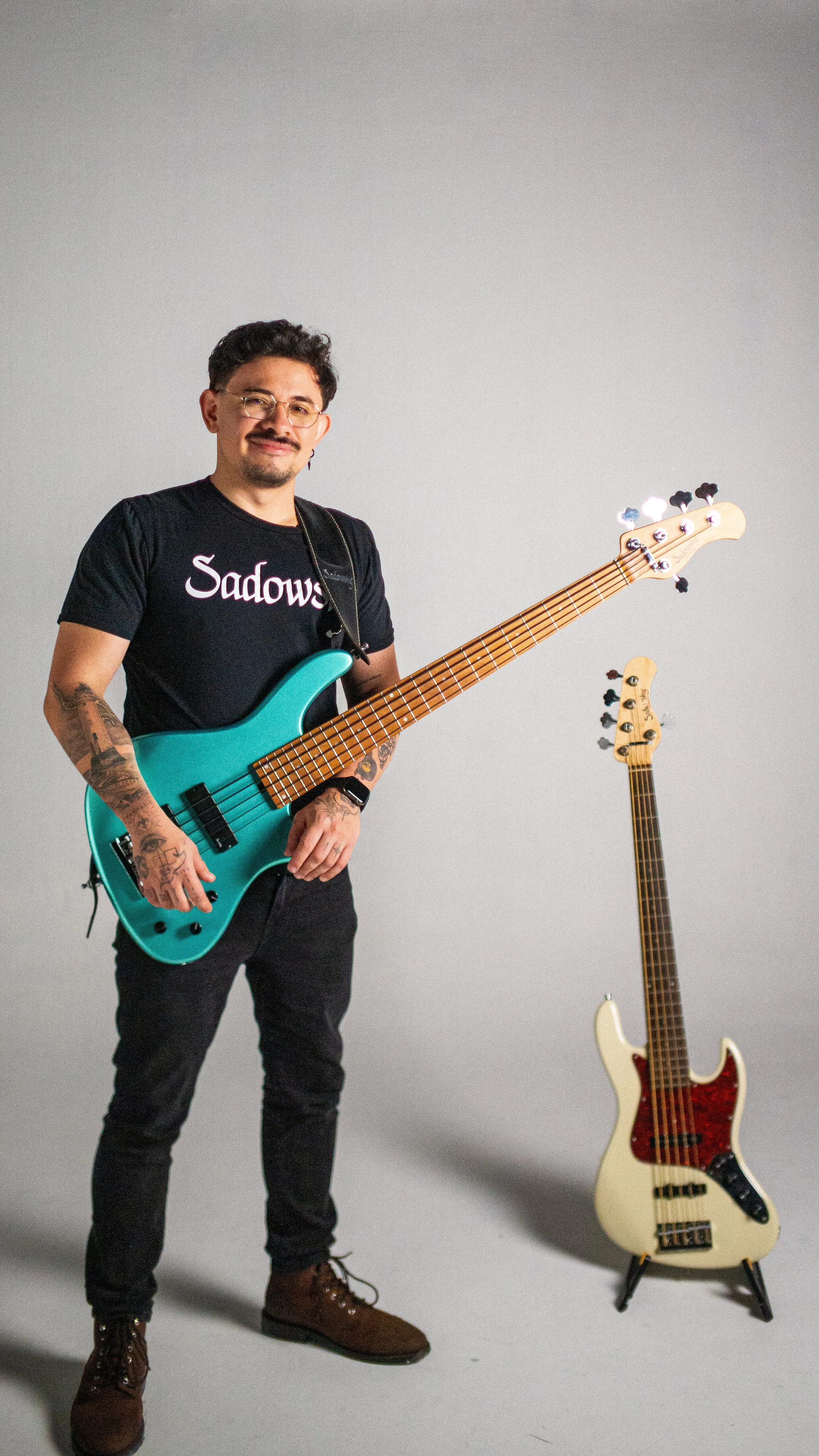 A man with glasses, tattoos, and a black 'Sadows' T-shirt holding a teal electric bass guitar, standing next to a white electric bass guitar on a stand against a plain gray background.