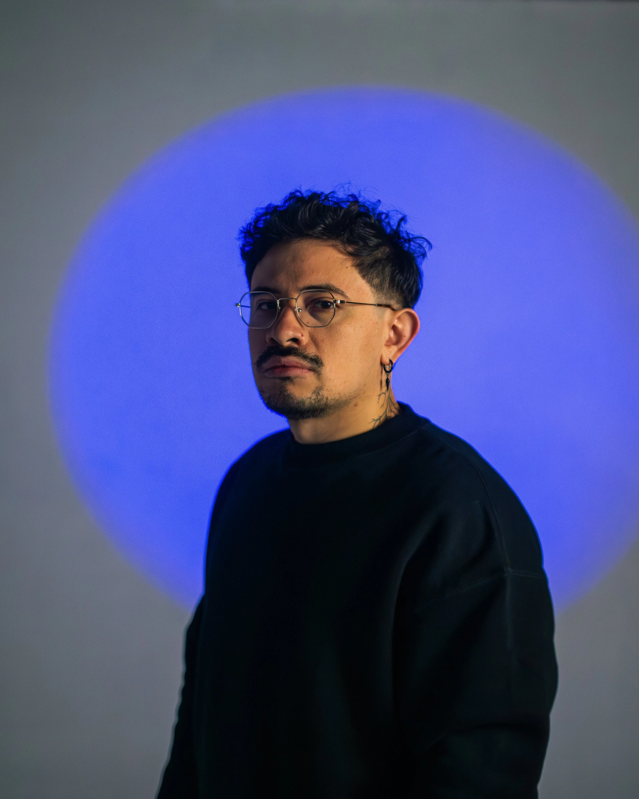 Portrait of a young man with dark hair and glasses, wearing a black shirt, standing against a background with a large glowing blue circle.
