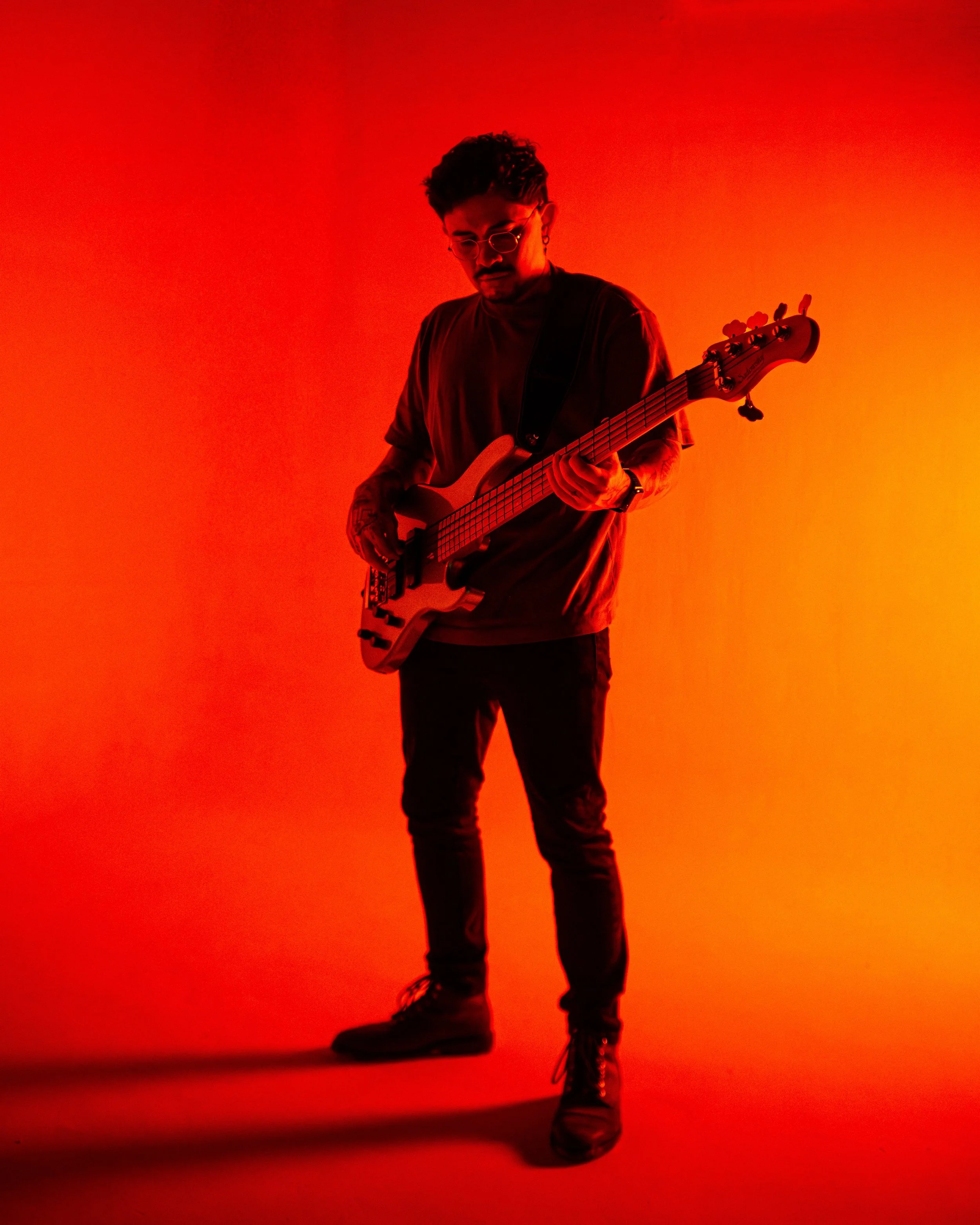 A person with glasses and tattoos playing an electric bass guitar under red-orange lighting, facing downward.