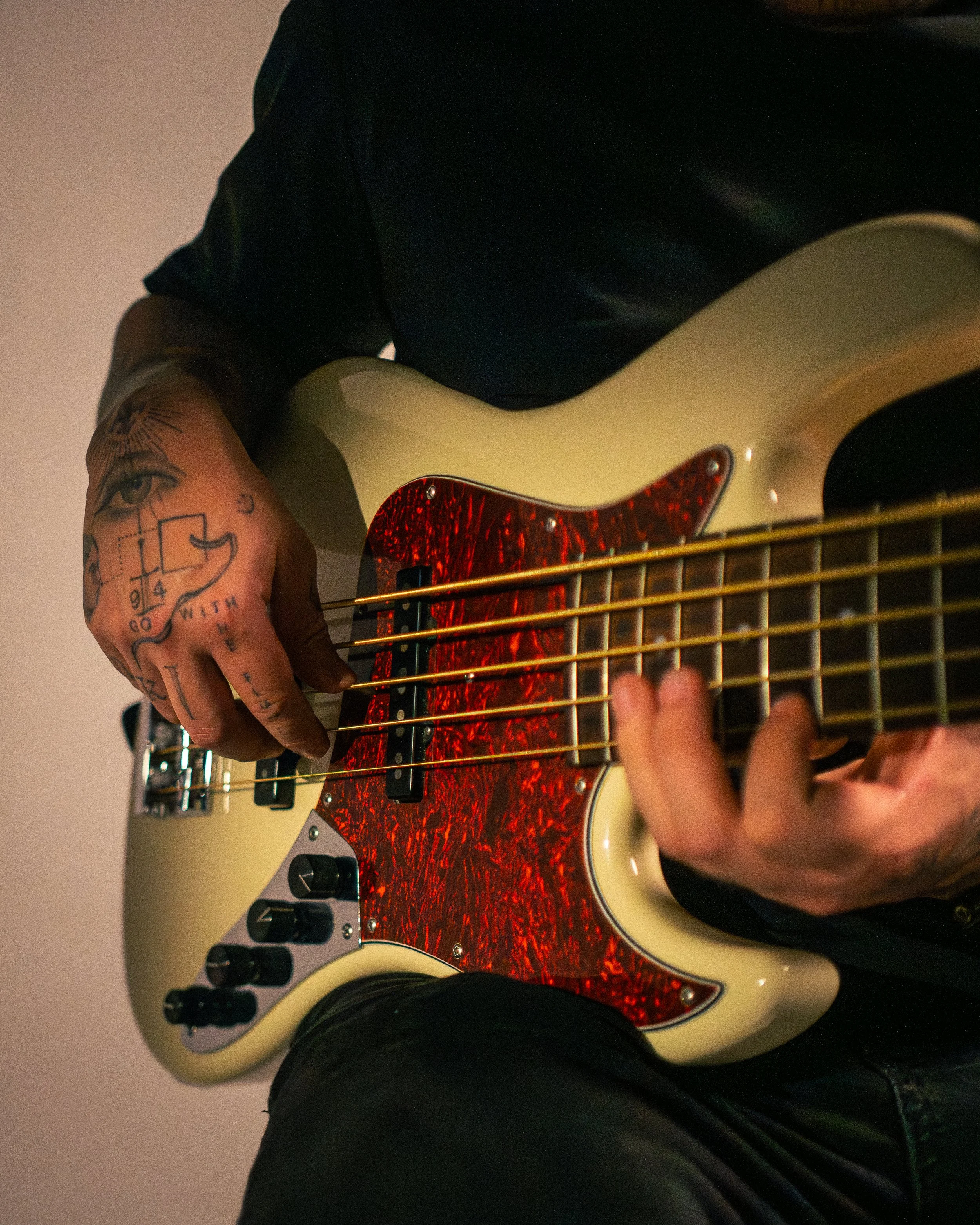 Close-up of a person playing an electric guitar with a cream-colored body, a red pickguard, and black control knobs, wearing a black long sleeve shirt and black pants.