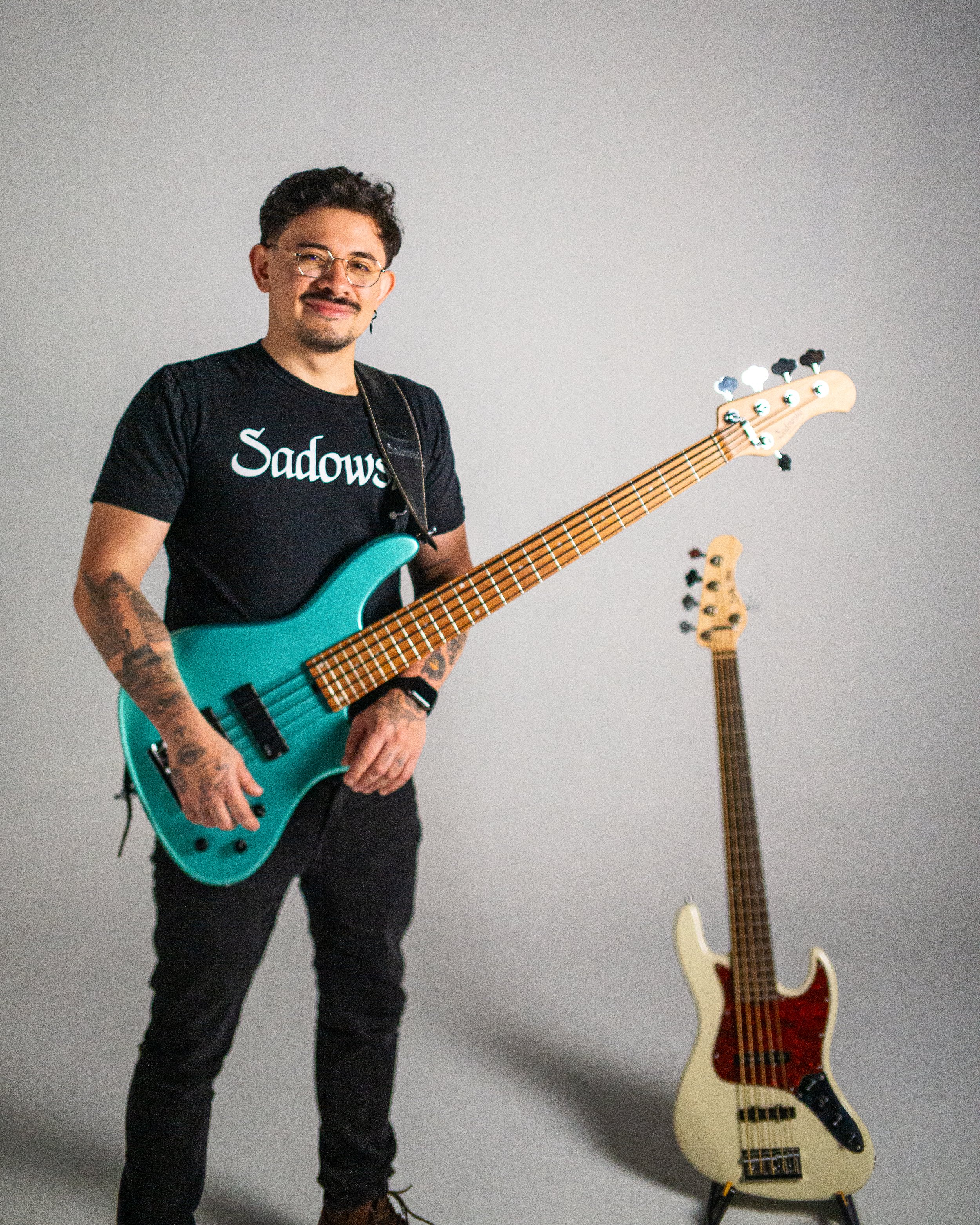 A man with tattoos, glasses, and a goatee holding a teal electric bass guitar, standing in front of a plain backdrop with another guitar visible in the background.
