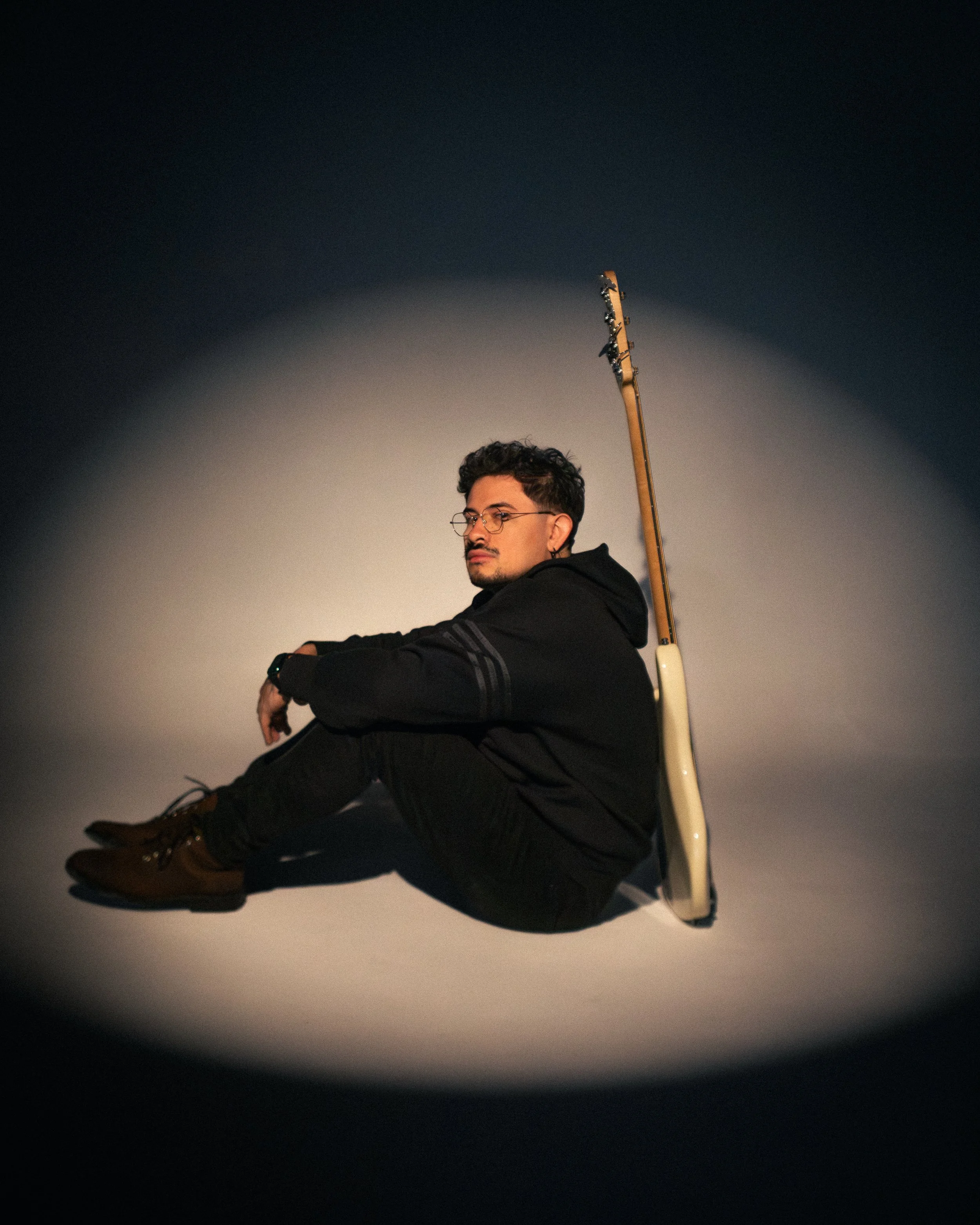 A man with glasses and curly hair sitting on the floor against a plain background, with a guitar resting on his shoulder.
