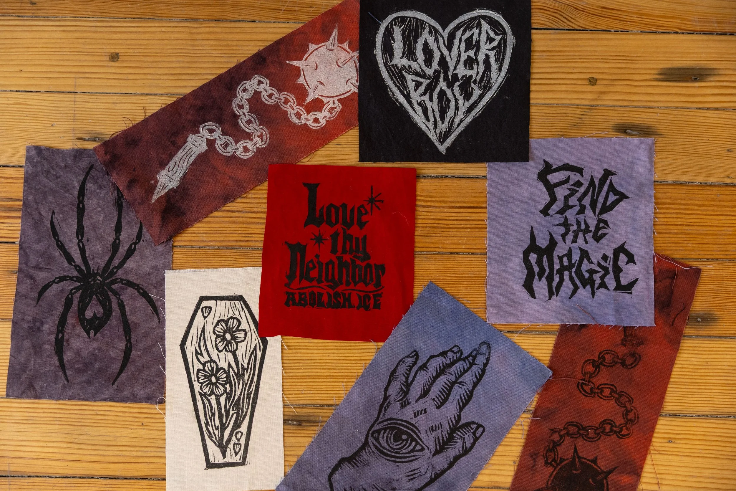 Collection of fabric patches with black and white tattoo-style designs, including a spider, coffin, heart, hand with eye, and chains, on a wooden surface.