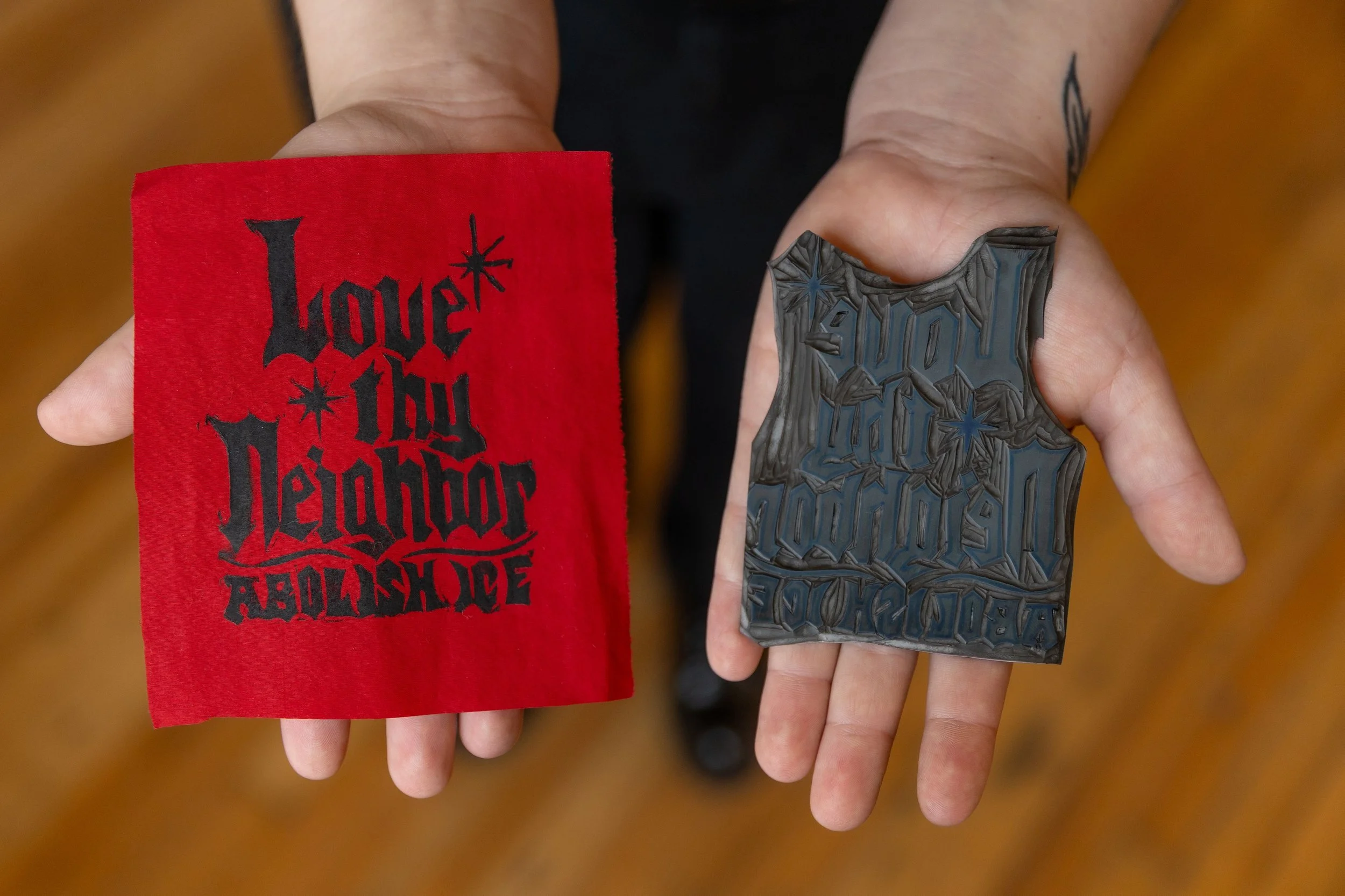 A person holding a red cloth with black text and a black rubber stamp with the same design. The cloth reads 'Love in the Heart of the Desert' in gothic font with stars. The rubber stamp has a reversed image of the text.