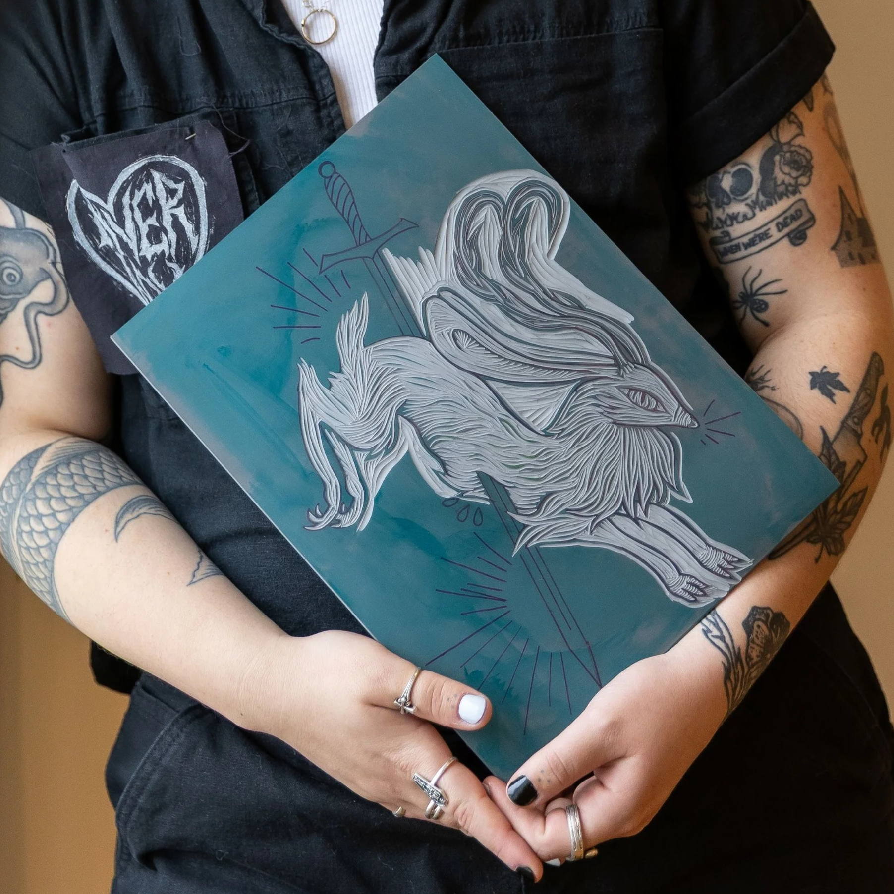 Person holding a graphic art print of a fox with a sword on a teal background, wearing a black shirt with patch and tattoos on arms.