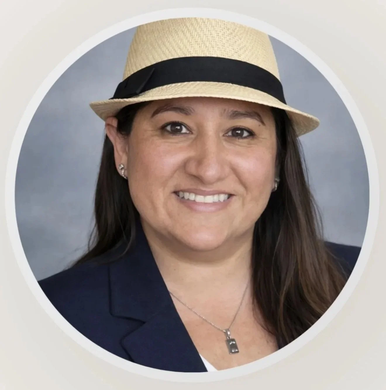 A woman wearing a straw hat with a black band, smiling, with long brown hair, earrings, and a necklace, posed against a neutral background.