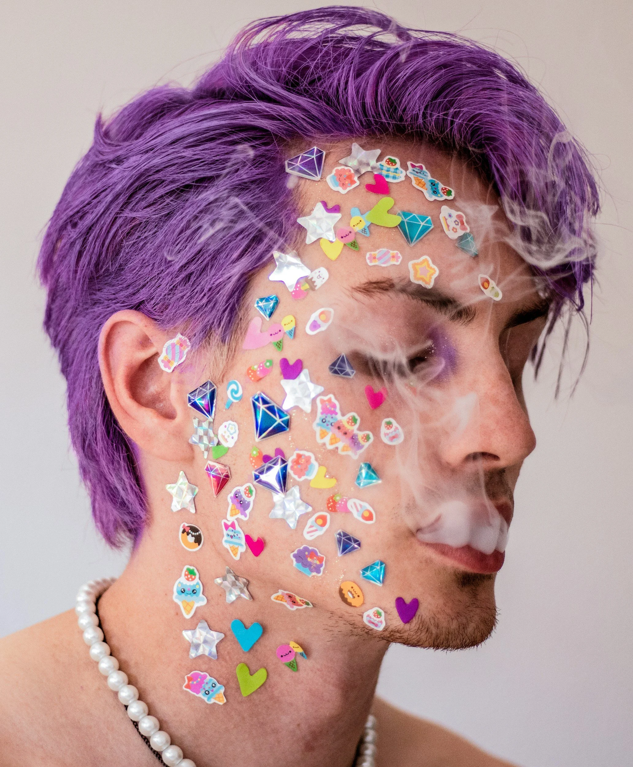 Junge mit lila Haaren trägt viele Glitzersticker und kleine Karten auf seinem Gesicht, trägt eine Perlenkette und raucht.