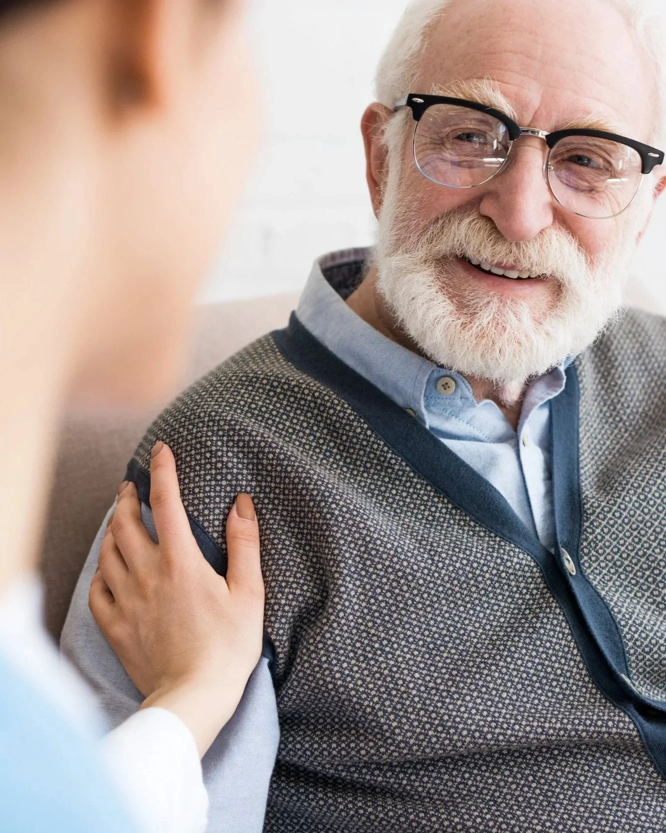 An elderly man with white hair, beard, and glasses smiling while having his shoulder held by a woman, possibly in a care setting.