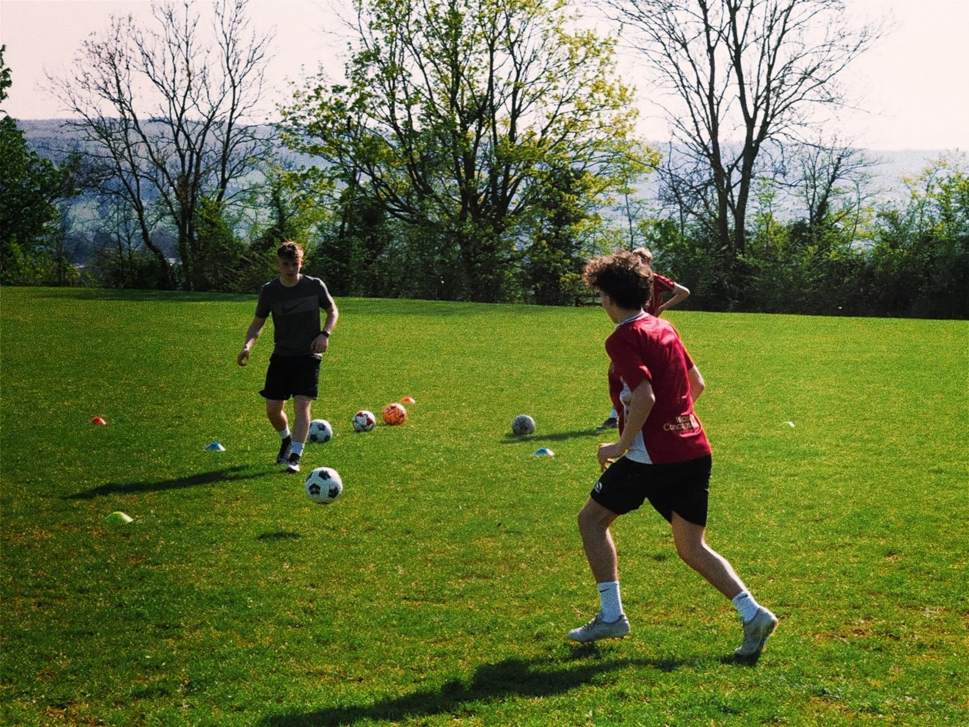 Three people playing football on a grassy field surrounded by trees on a sunny day.
