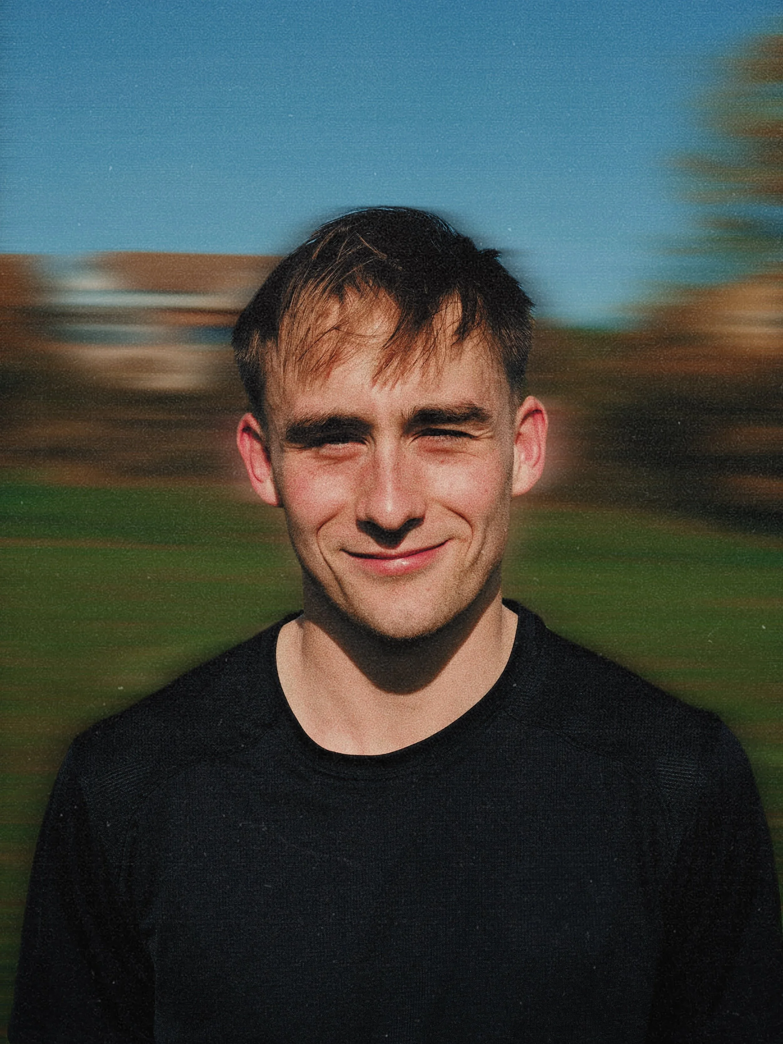 A young man with dark hair smiling outdoors on a sunny day.