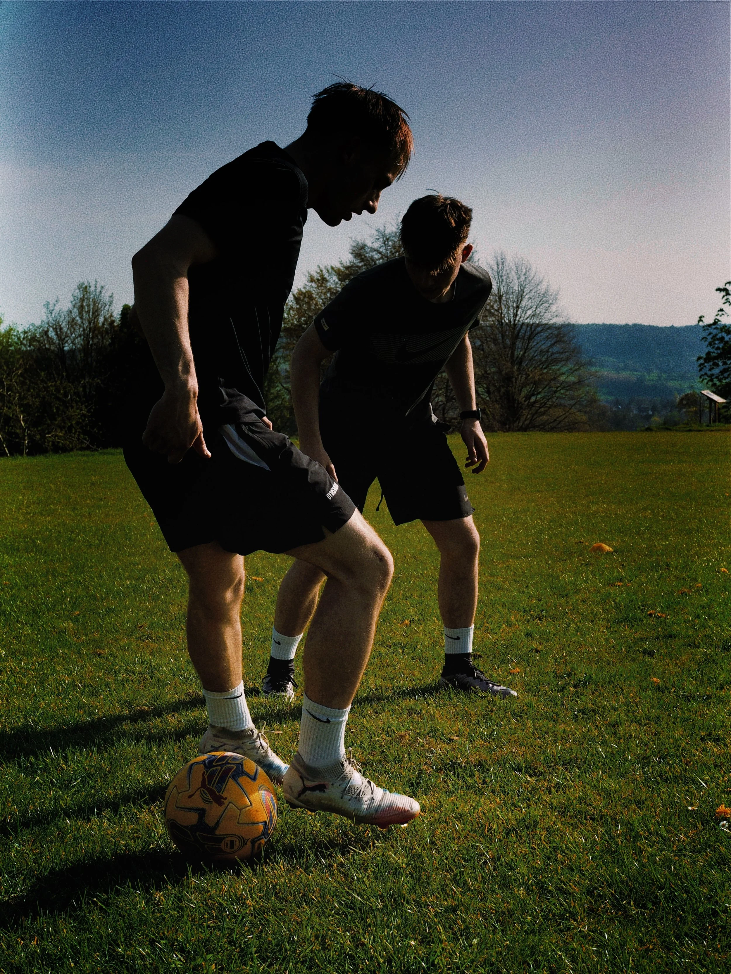 Two men iplaying football outdoors on a grassy field during late afternoon or evening. The scene has a scenic backdrop with trees and distant hills under a clear sky.