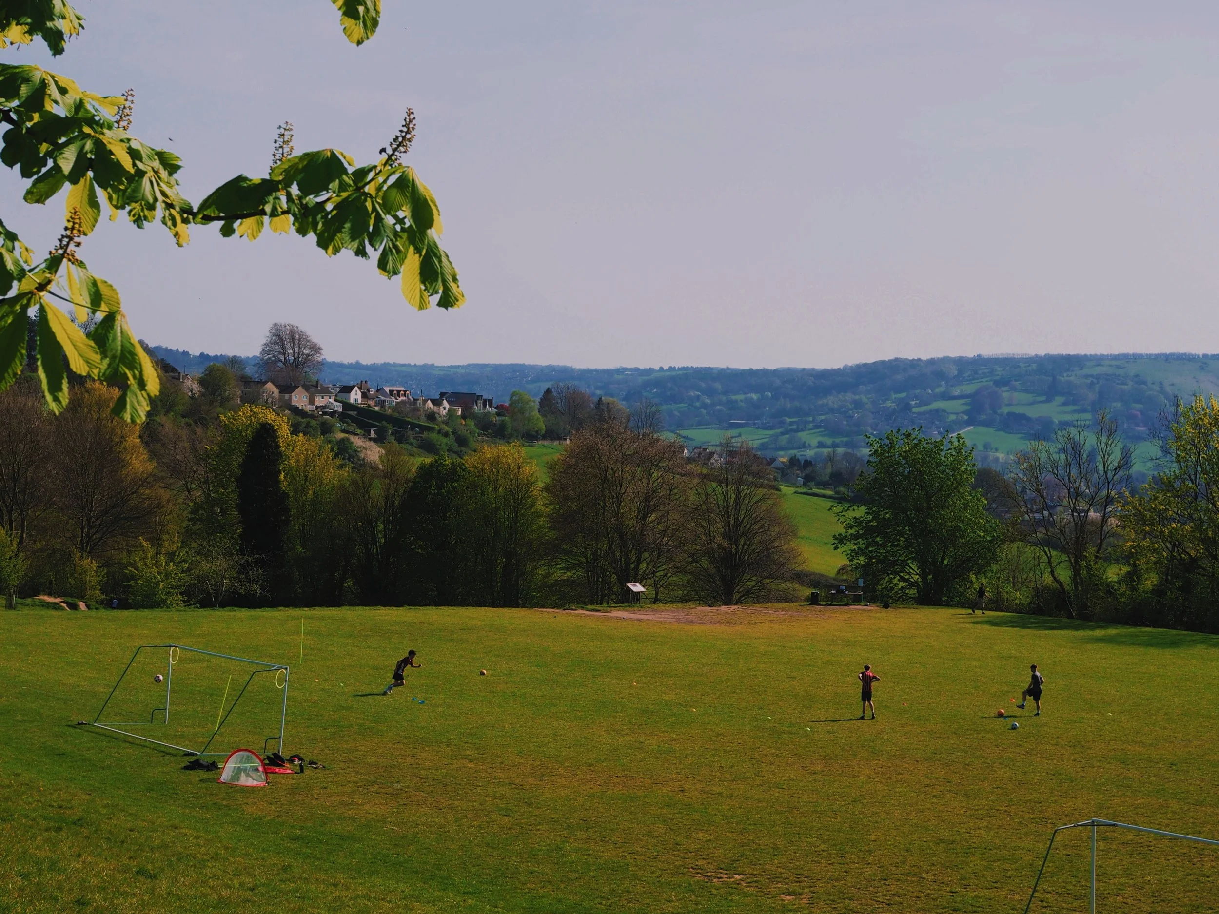Three people playing football on a grassy field with trees and hills in the background.