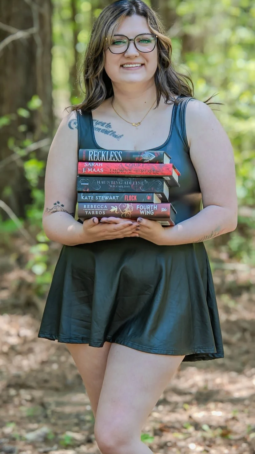 A woman with glasses and tattoos, wearing a black dress, standing outdoors in a forest, holding a stack of seven books. She is smiling.