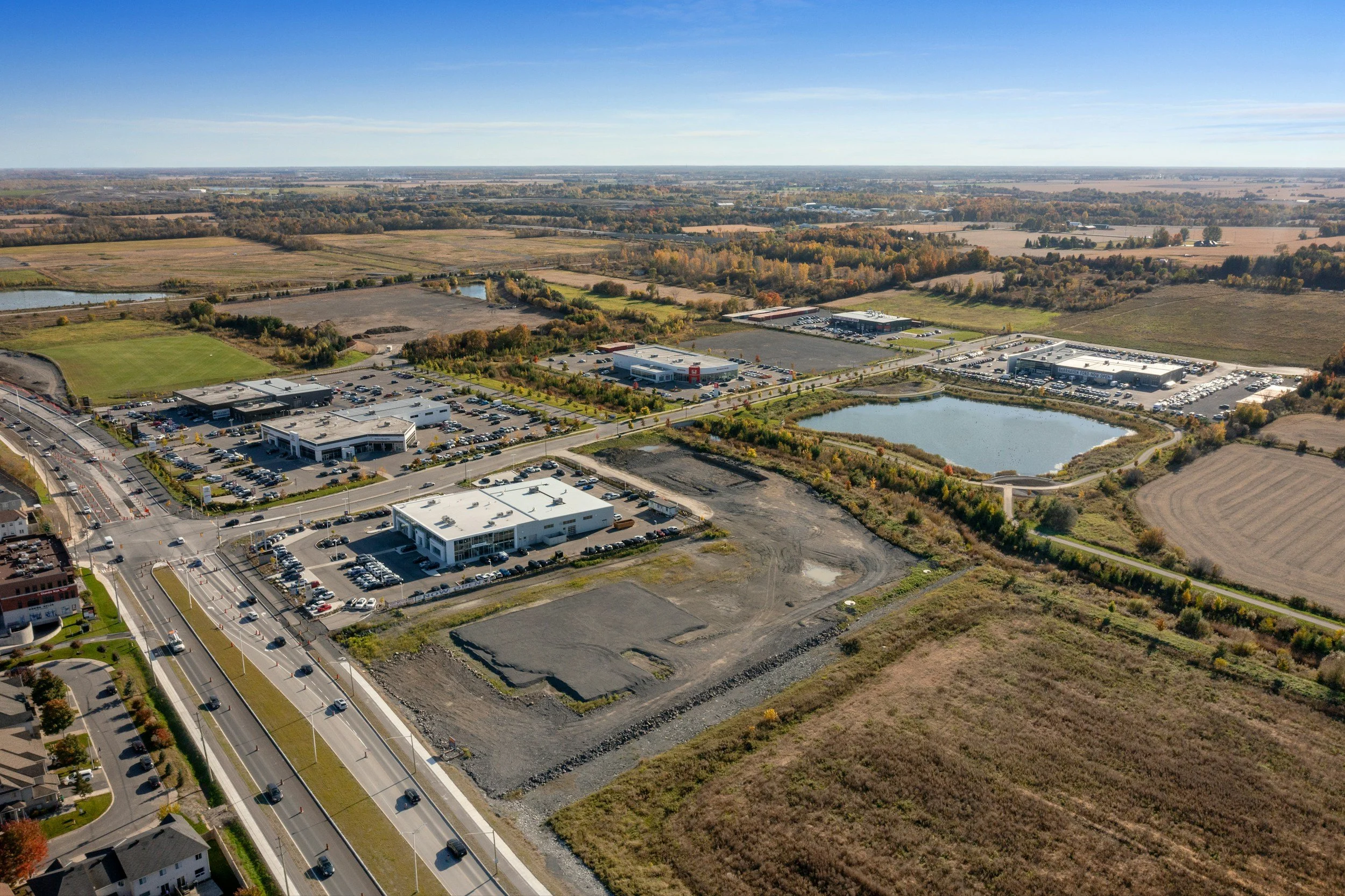 Aerial view of a suburban area with commercial buildings, parking lots, a small pond, and fields with trees and farmland in the background under a clear blue sky.