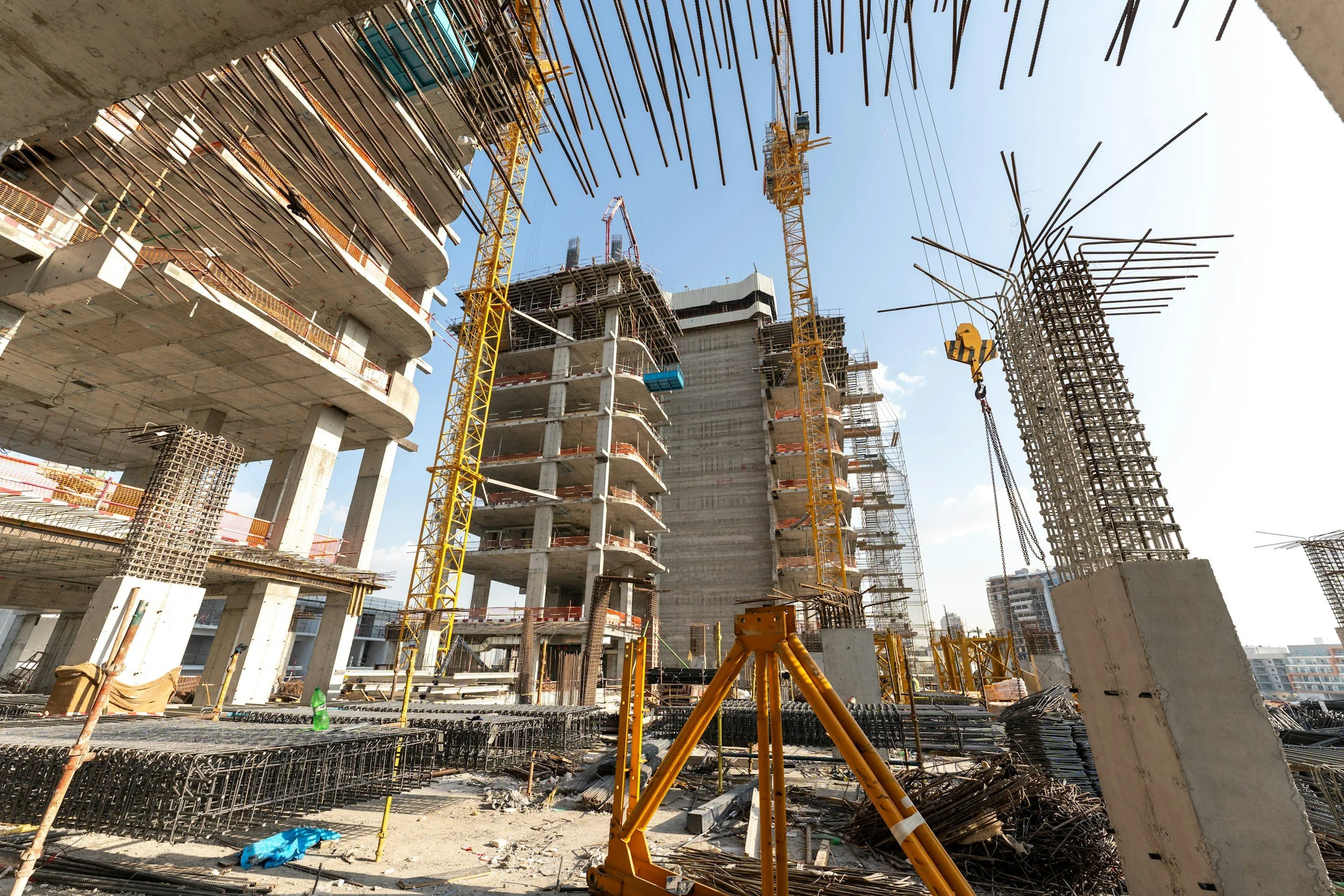 Construction site with partially built high-rise buildings, yellow cranes, and exposed rebar, under a clear blue sky.