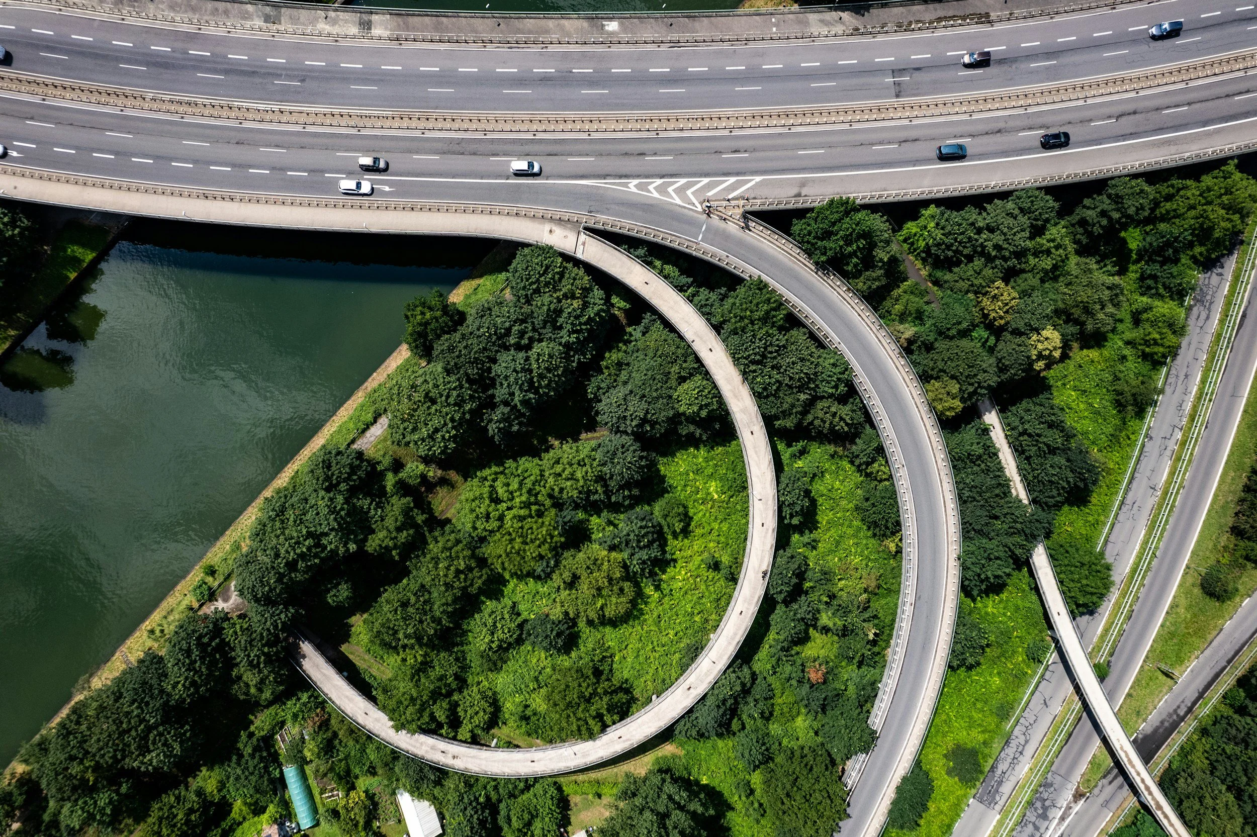An aerial view of a multi-level highway system with several vehicles traveling on the roads surrounded by green trees and a body of water.
