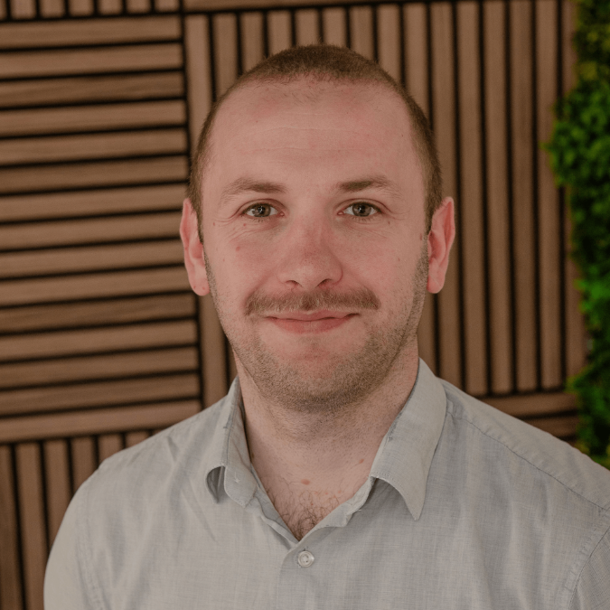 Cameron Berry smiling and standing indoors with a wooden fence and greenery in the background.