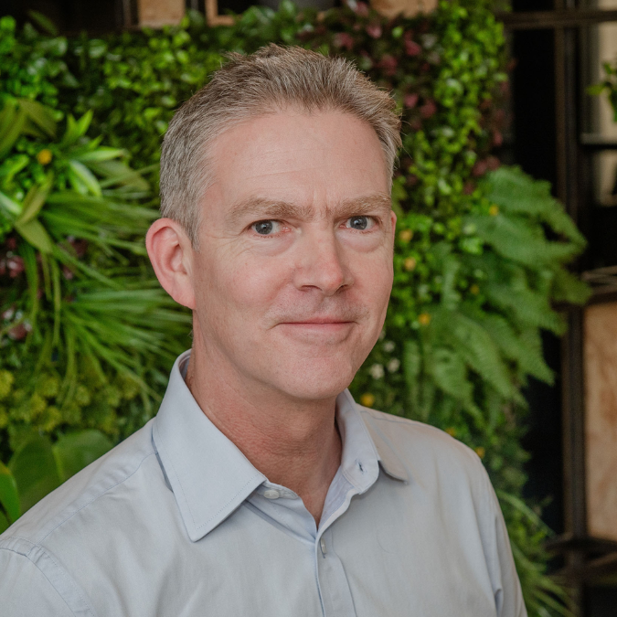 John Hinett standing indoors with green foliage and plants in the background.