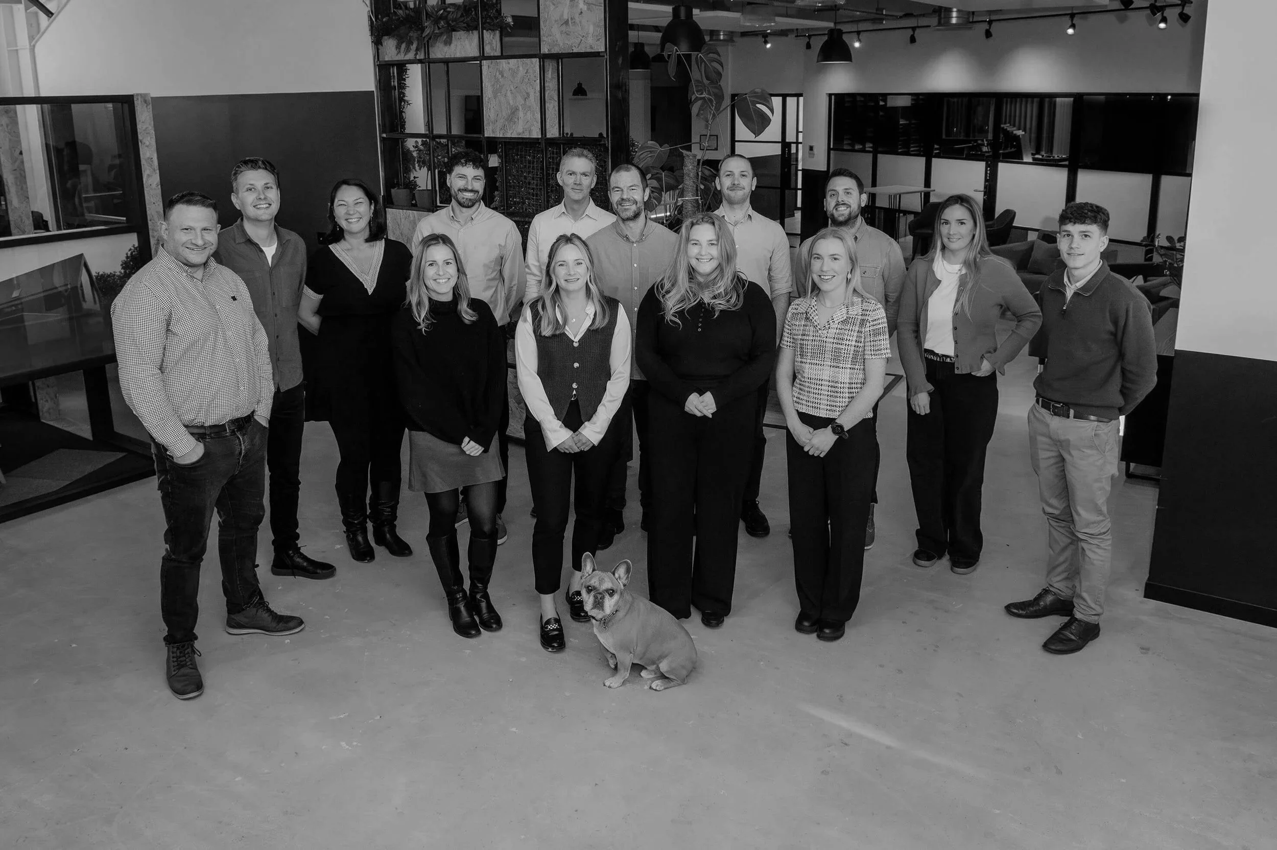 Group of fifteen people and a dog posing for a photo in an indoor office space, with some smiling and others looking at the camera.