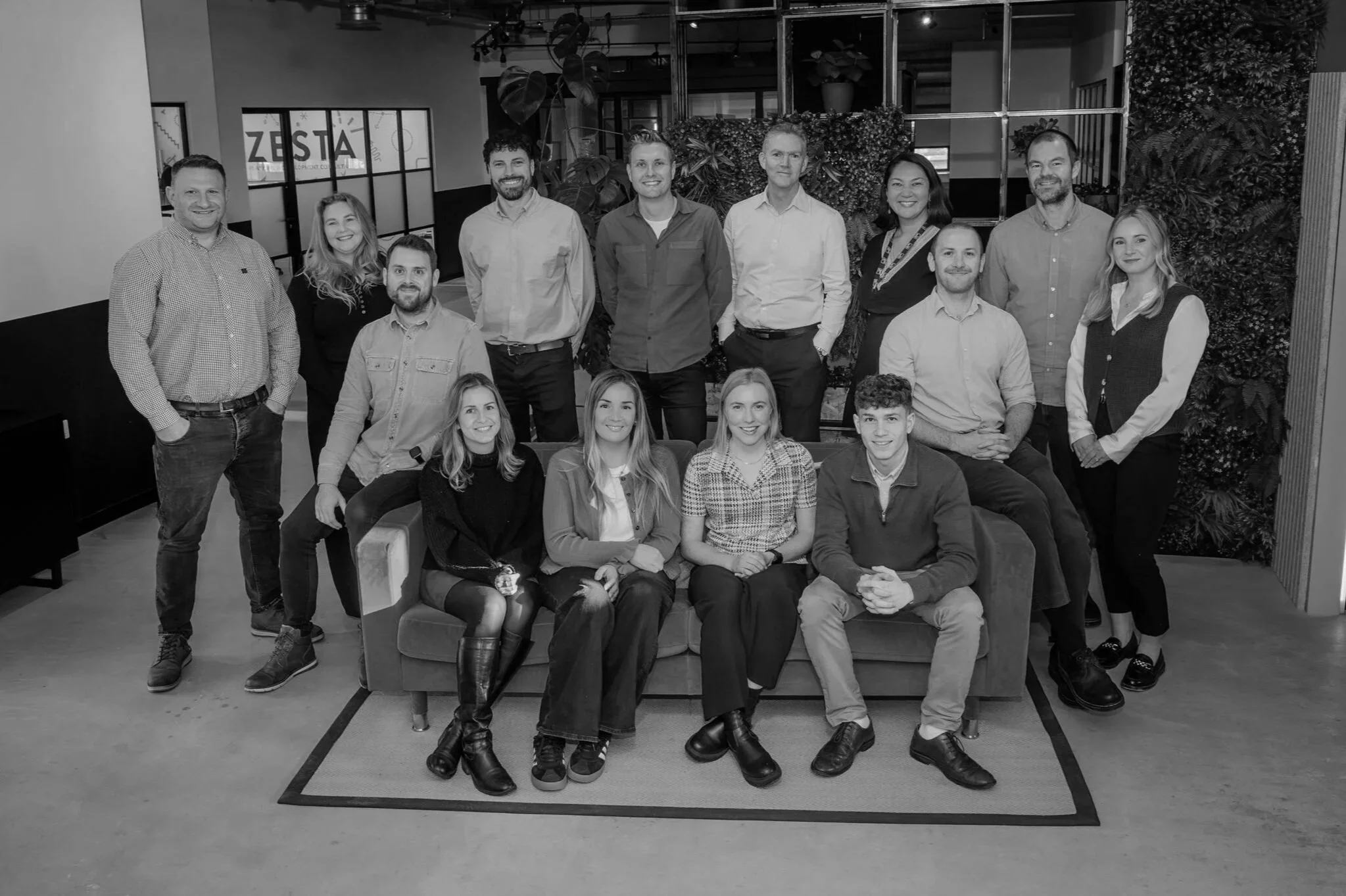 Group photo of 13 coworkers in a Zesta Planning office, some sitting on a sofa and others standing behind, with plants and office decor in the background.