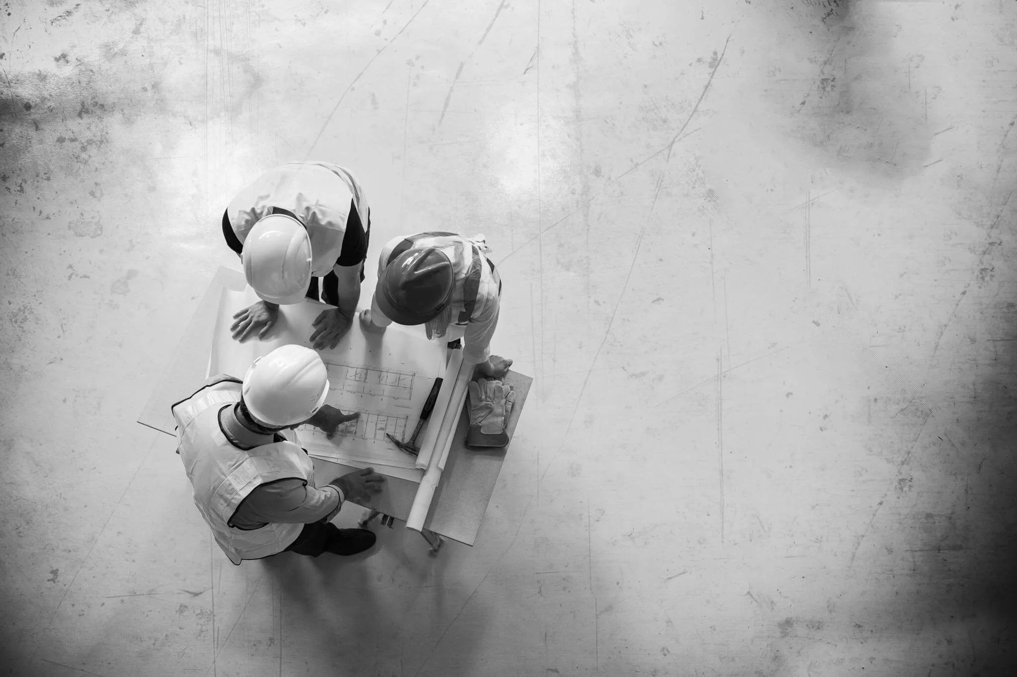 Top-down view of three construction workers wearing white hard hats and safety vests gathered around a table with blueprints and plans, on a plain, textured floor.
