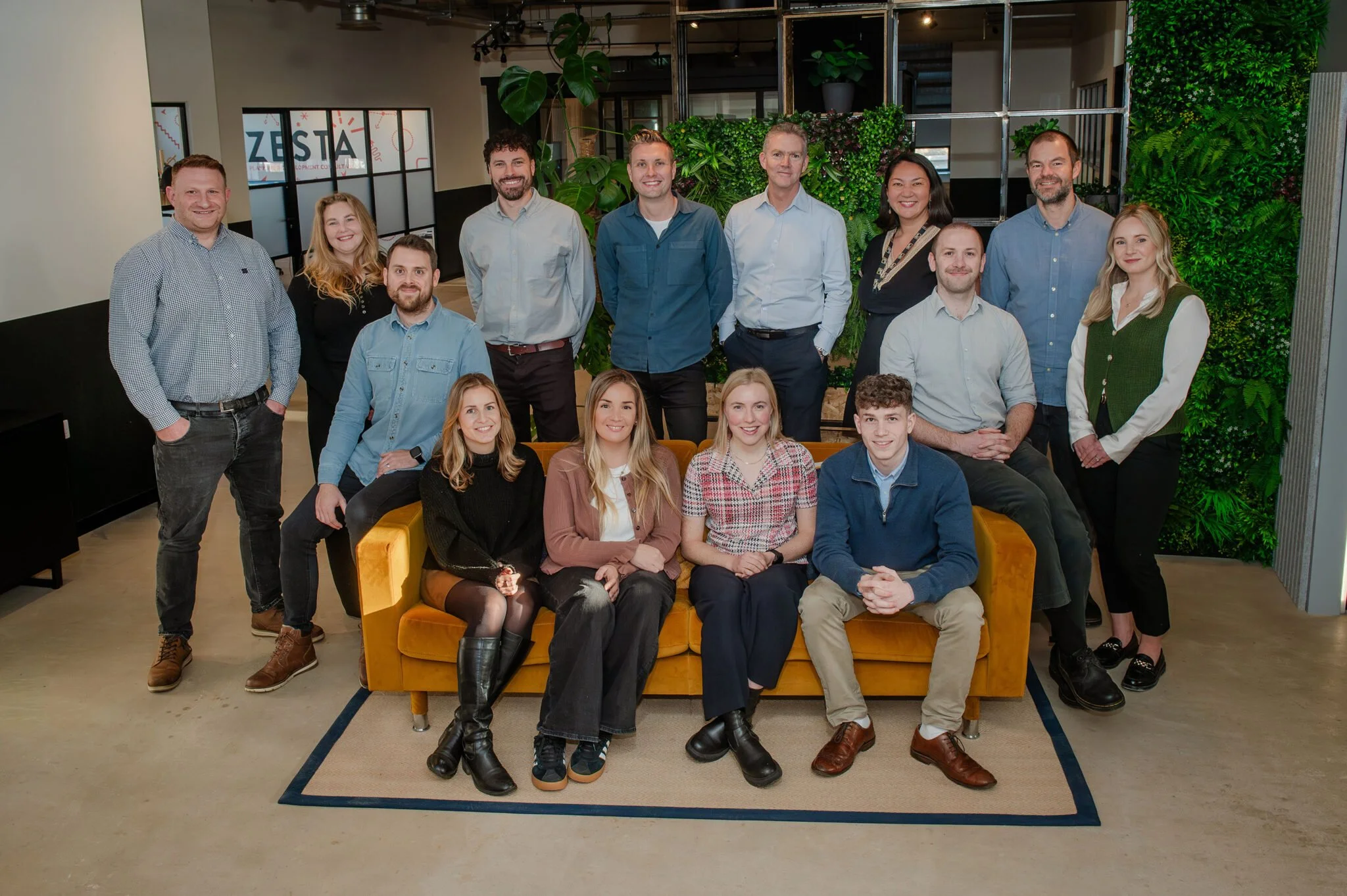 A group of fifteen young adults posing together for a photo in a Zesta Planning modern office setting, with some sitting on a yellow sofa and others standing behind it, surrounded by green plants.