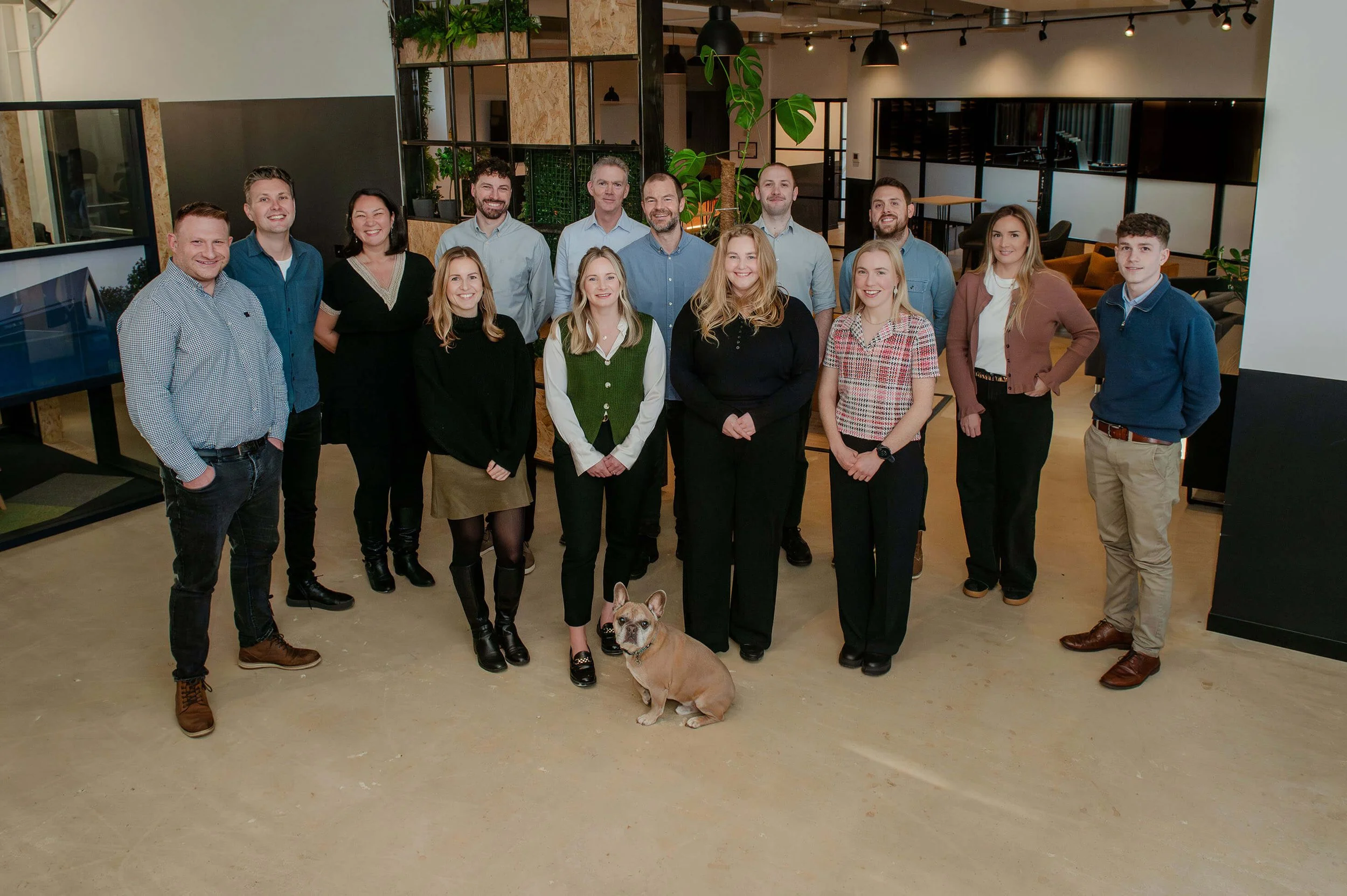 A group of fifteen people posing for a photo in an indoor modern Zesta Planning office space, with some smiling and standing in front of a large potted plant, accompanied by a French Bulldog dog sitting on the floor.