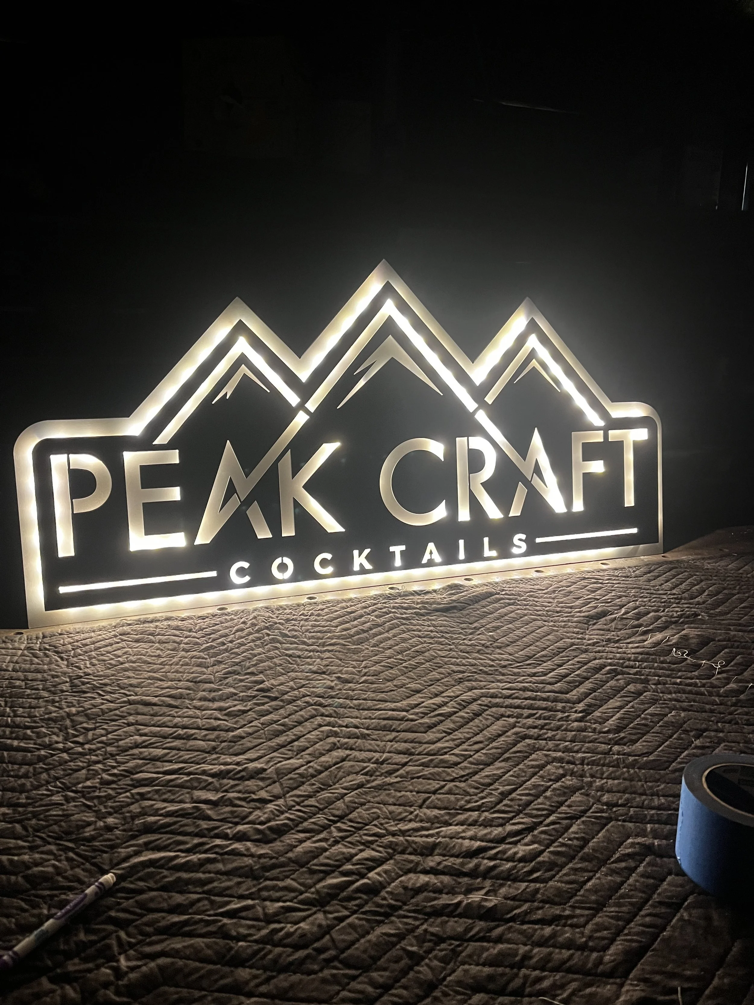 Backlit sign with mountain outline and the text 'Peak Craft Cocktails' on a textured surface