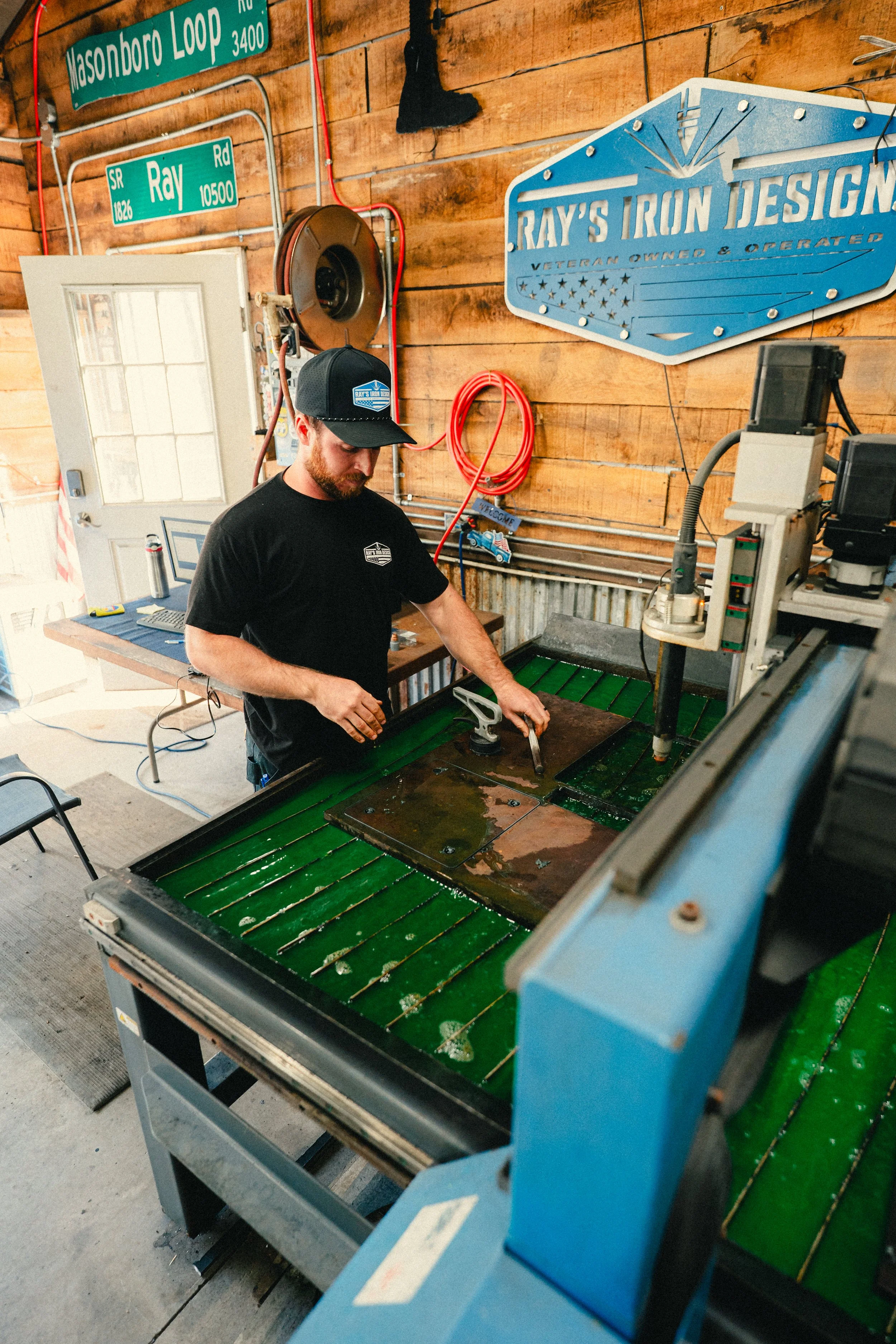 A man operating a CNC machine with a green work surface in a woodworking shop, with wood wall decor and signs in the background.