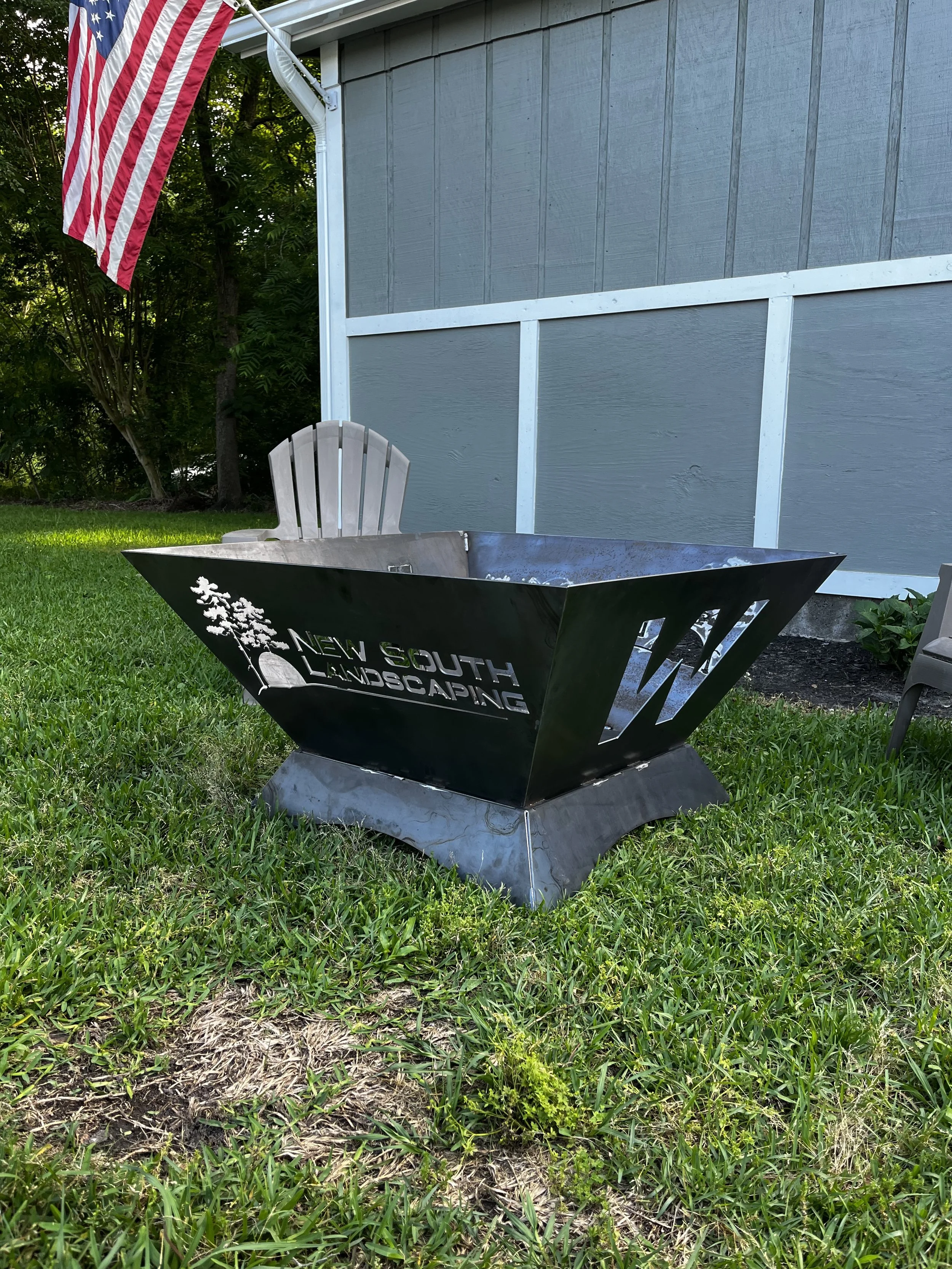 Black metal fire pit with the logo and name 'New South Landscaping' on the side, set on a grassy yard next to a gray house with blue trim, an Adirondack chair, and an American flag.