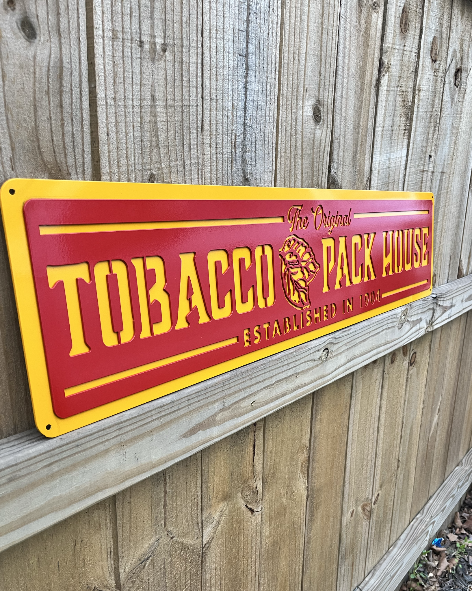 Red and yellow sign for Tobacco Pack House with the text and logo, mounted on a wooden fence.