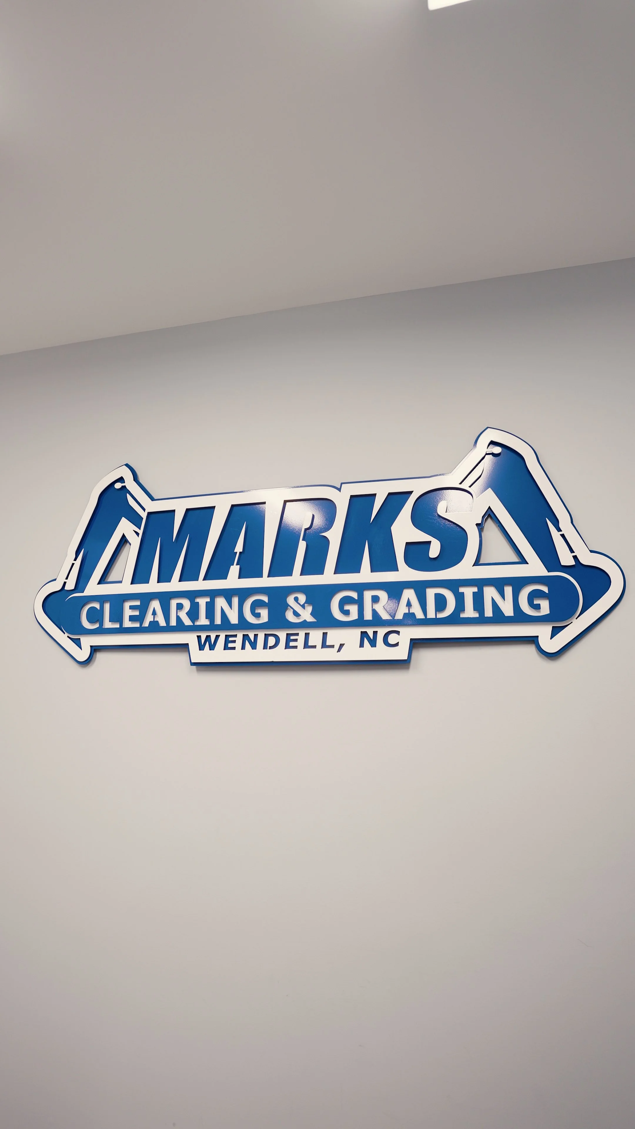 Blue and white sign for AMARKS Clearing & Grading in Wendell, North Carolina, mounted on a plain white wall.