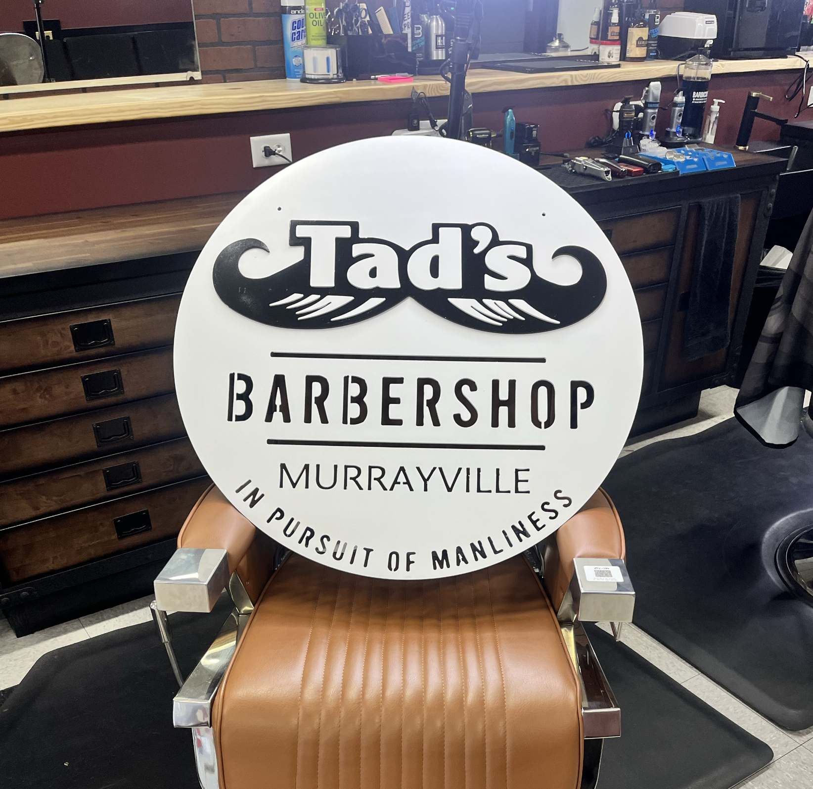 A circular sign for Tad's Barbershop in Murrayville, placed on a salon chair inside a barber shop. The sign has black and white text and graphics, and the shop interior features a wood counter, barber tools, and other hair care products.