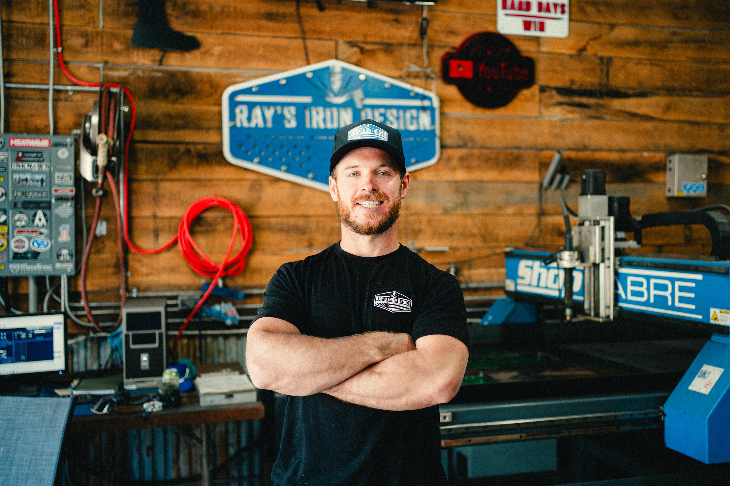 A man with a beard and a black cap, smiling with arms crossed, standing in a woodworking or metalworking workshop with tools, machine, and signs on wooden wall.