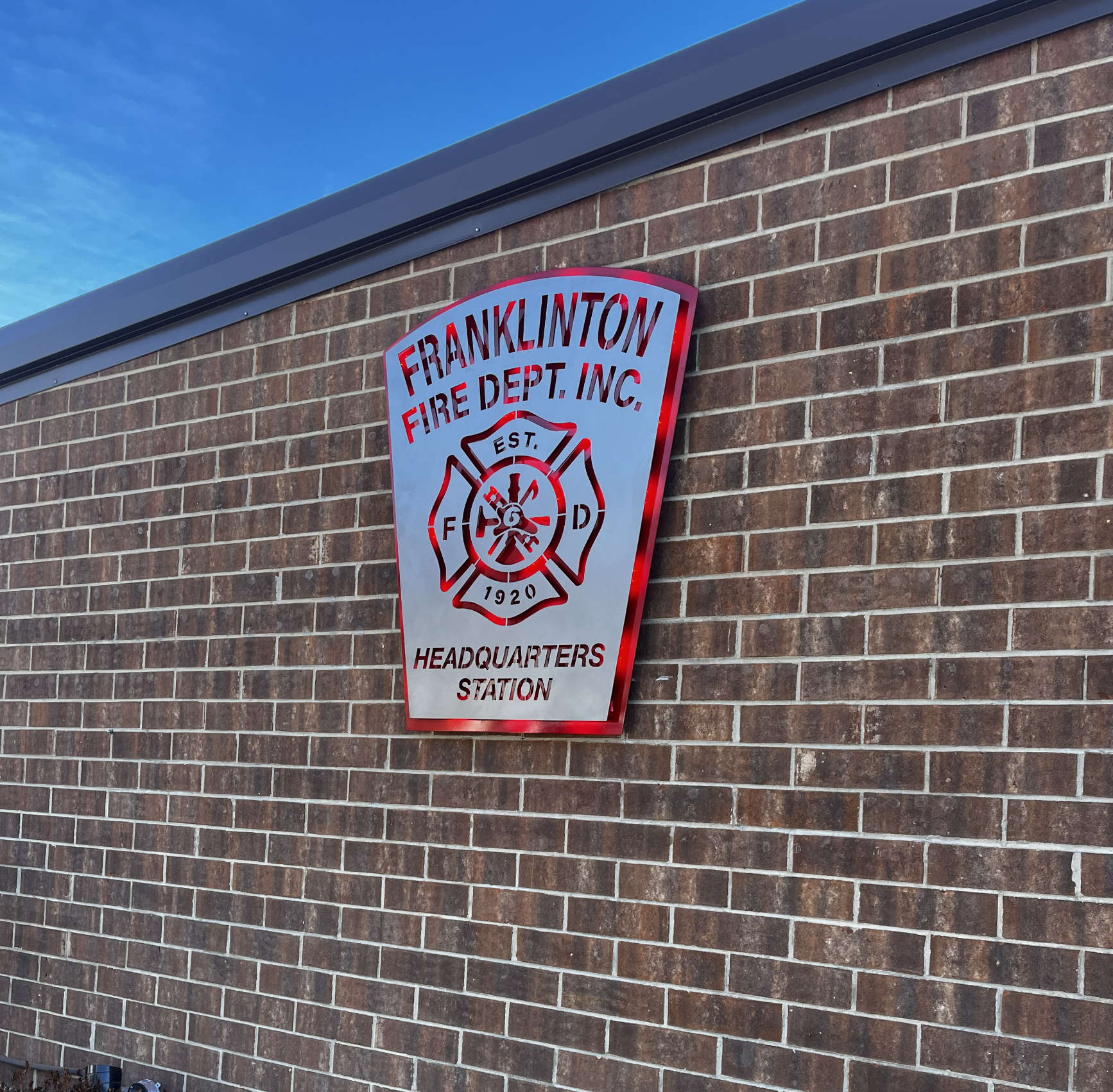 A metal sign on a brick wall that reads 'Franklinton Fire Dept. Inc. Headquarters Station', featuring a firefighter's emblem with a ladder, axe, and hose, with the establishment year 1920.