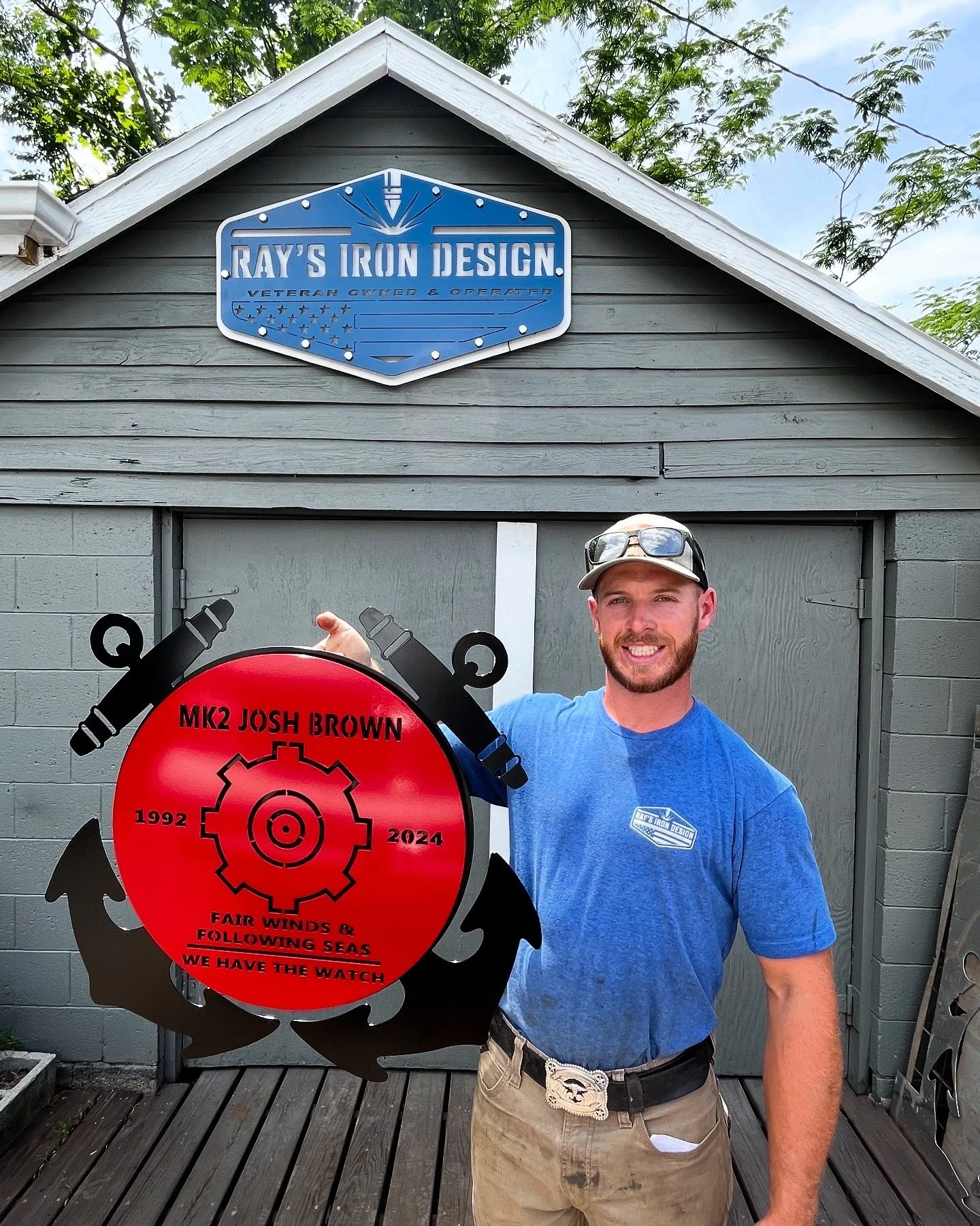 A man in a blue T-shirt and tan pants holding a red sign with black gear and shark fin designs in front of a gray building with a sign that reads 'Ray's Iron Design'.