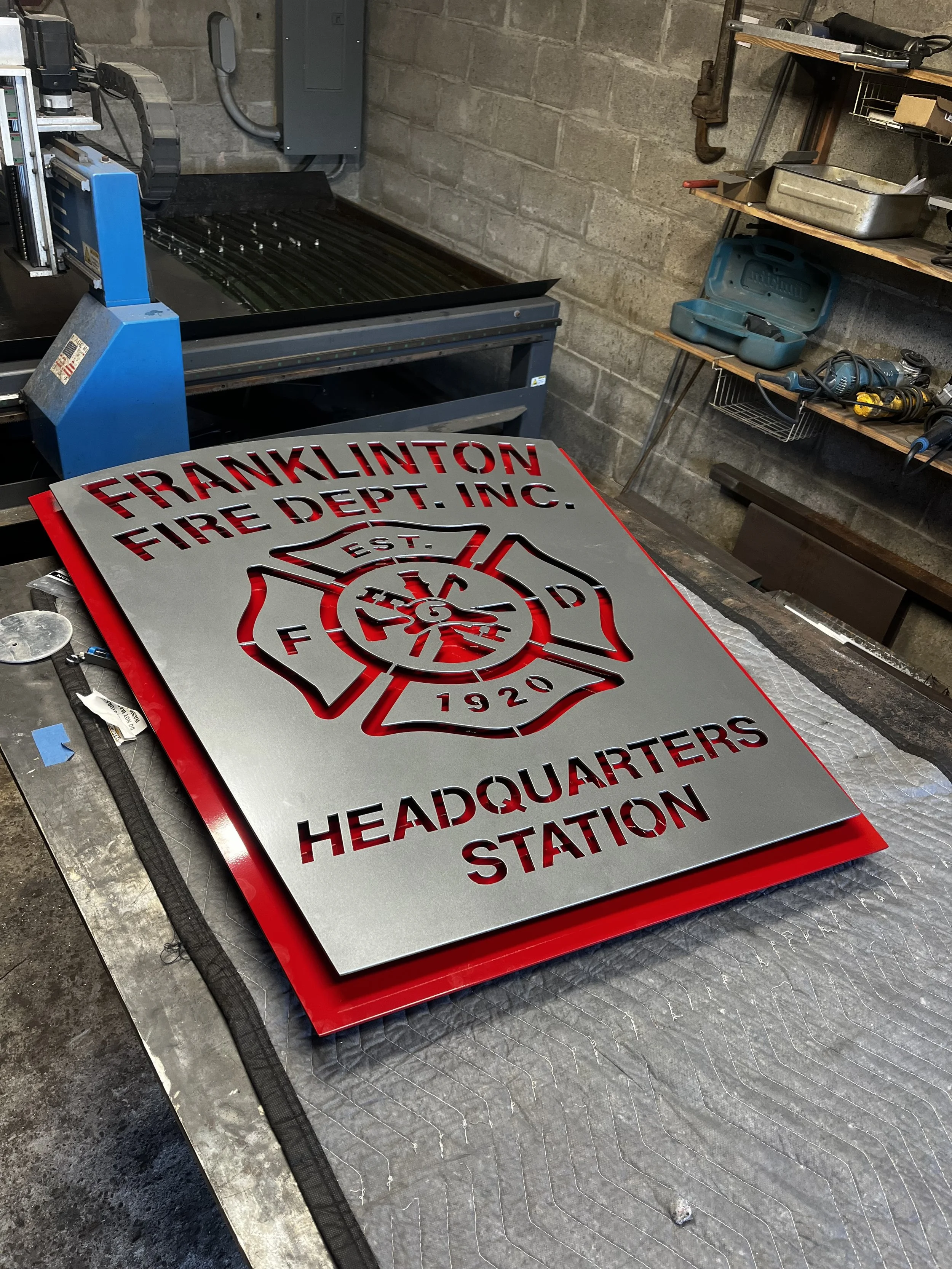 A metal sign for the Franklin Fire Department Inc. Headquarters Station, featuring a red border and a fire department emblem, placed on a workbench in a workshop.