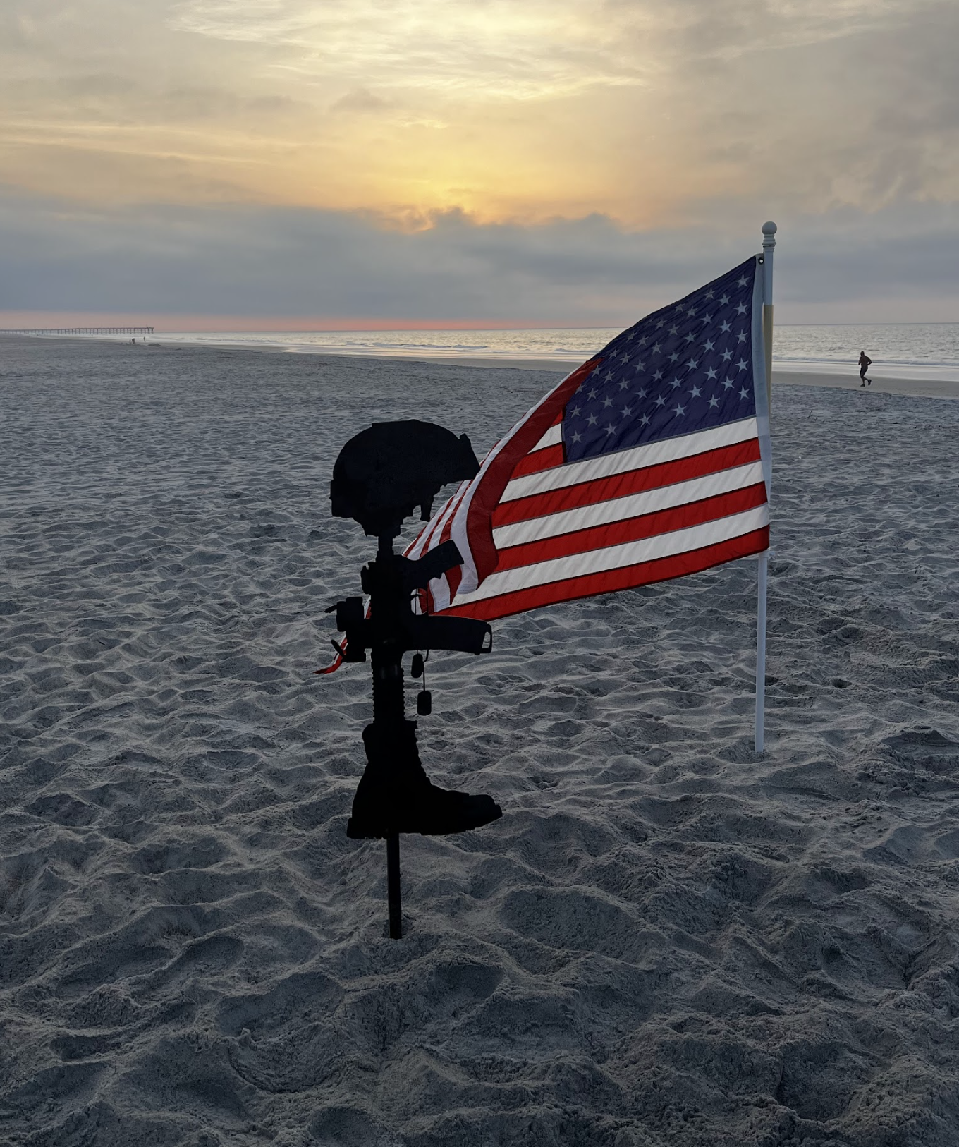 American flag planted in the sandy beach with a sunset view over the ocean, and a person walking in the distance.