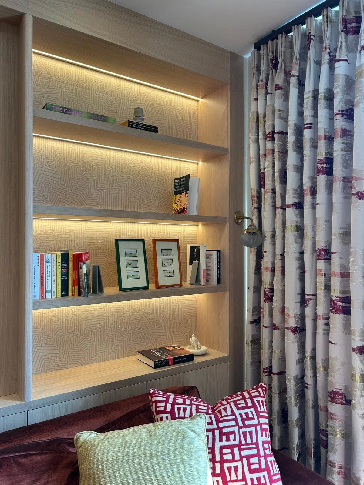 A wooden bookshelf with backlighting, containing books, framed pictures, and decorative objects, adjacent to patterned curtains and pillows on a sofa.