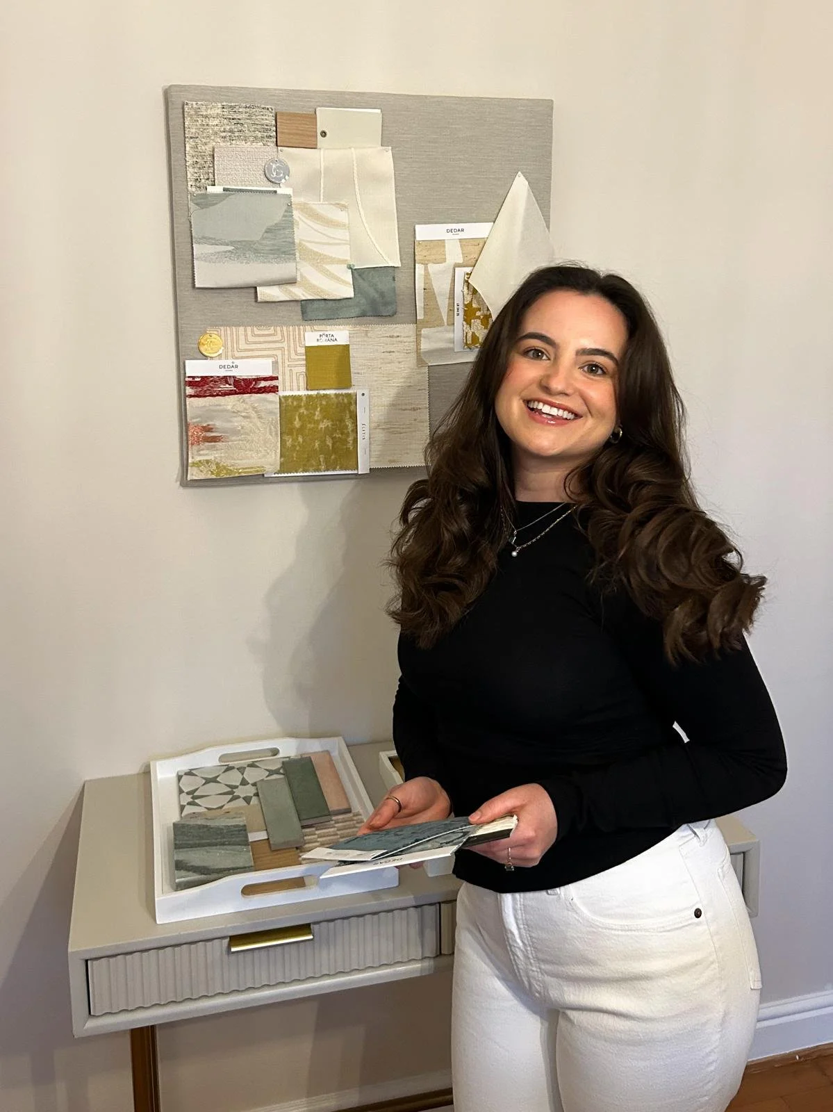 A woman with long brown hair, wearing a black top and white pants, is standing indoors near a small white table. She is holding a sample book, and behind her, there is a wall-mounted fabric and paint sample display, along with various fabric swatches on the table.