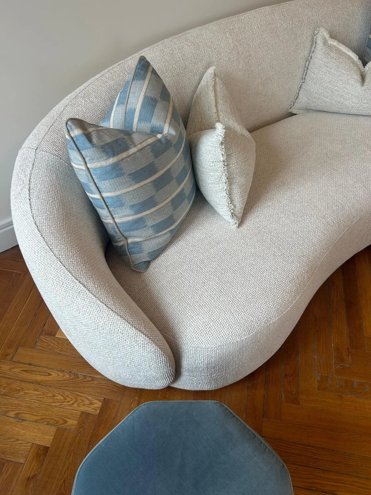 A curved beige upholstered sofa with three decorative pillows, including one blue and white striped pillow and two solid beige pillows, on a wooden floor.
