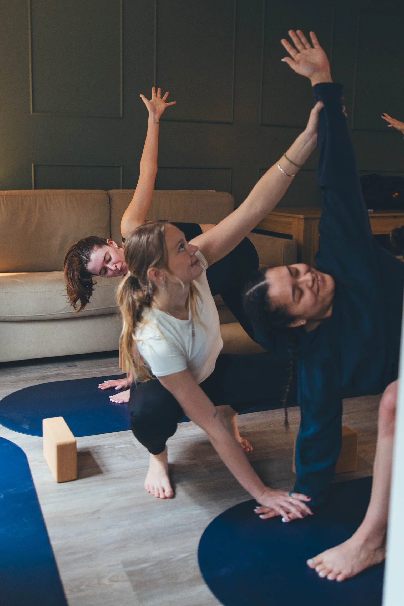 Four women participating in a yoga session in their airbnb. They are on yoga mats, practicing a pose while supporting each other, with a couch and dark wall panels in the background. Private Yoga session in their accomodation in Banff, Canmore.