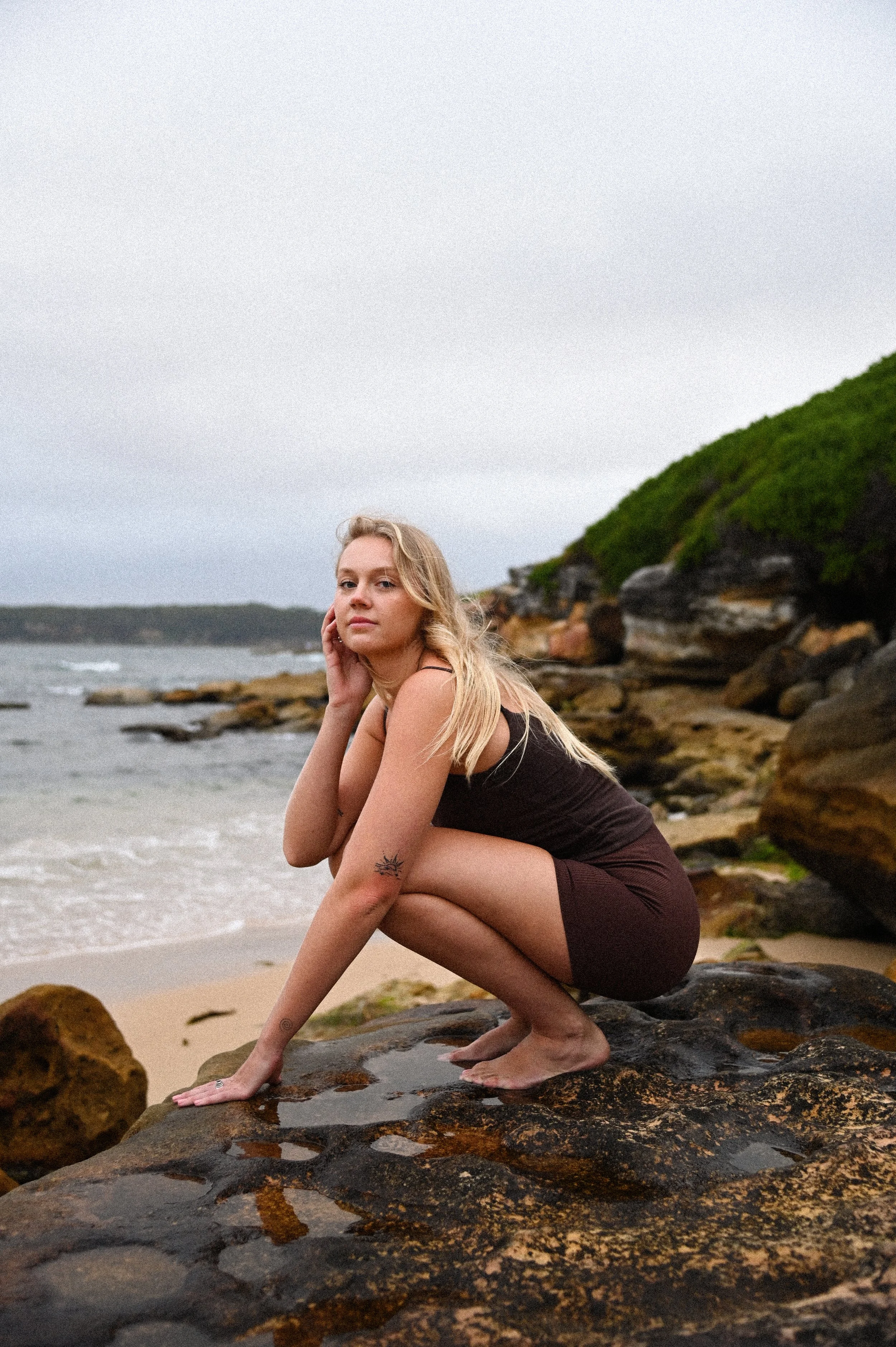 A young woman with long blonde hair and tattoos on her arm, squatting on rocks at the beach, resting her chin on her hand and looking at the camera, with a background of ocean waves, sandy shore, rocky cliff, and cloudy sky.