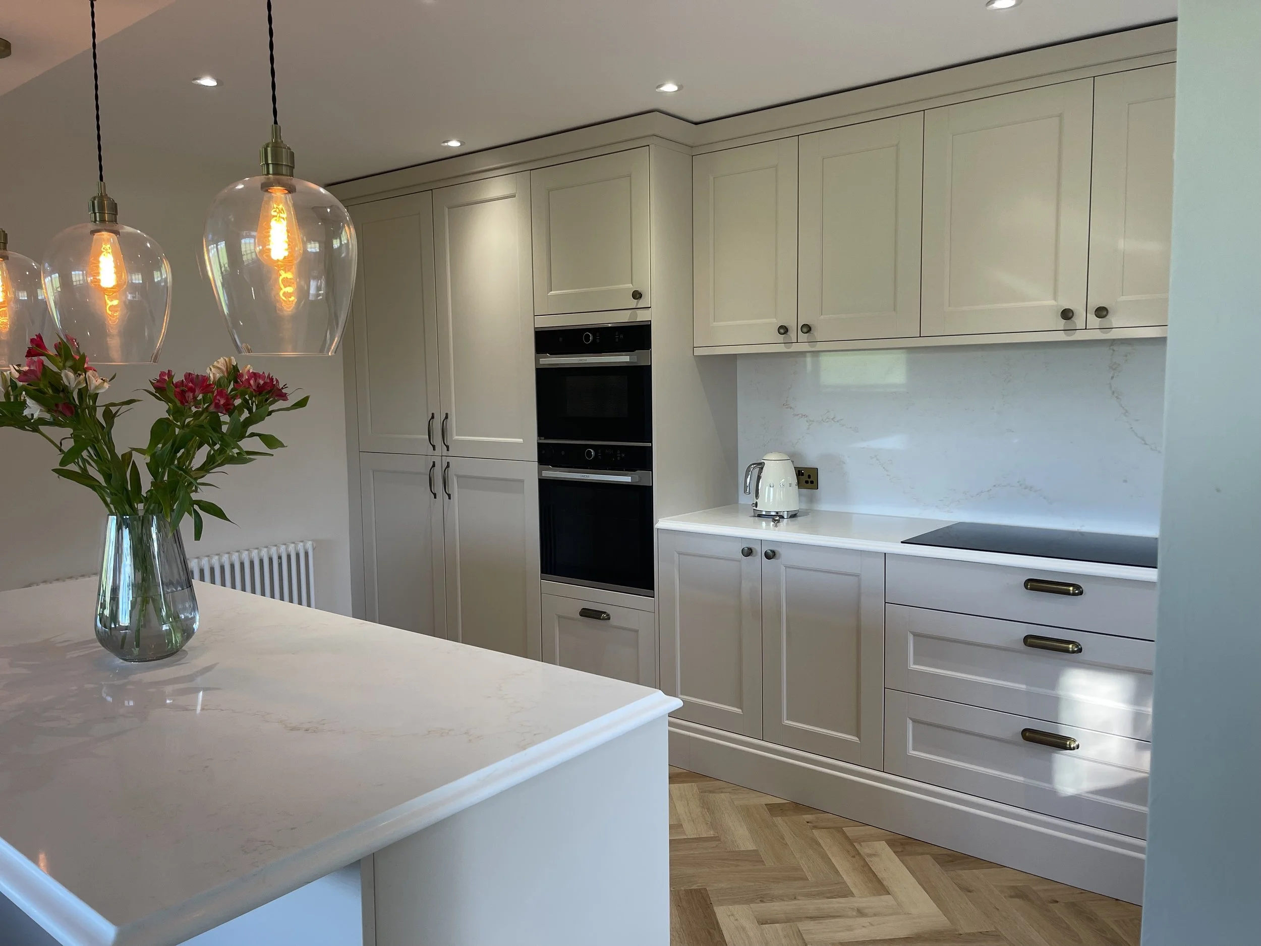 Modern kitchen with white cabinets, marble backsplash, black appliances, a white kettle, and a vase with pink flowers on a white marble countertop. Four pendant lights hang above the counter.
