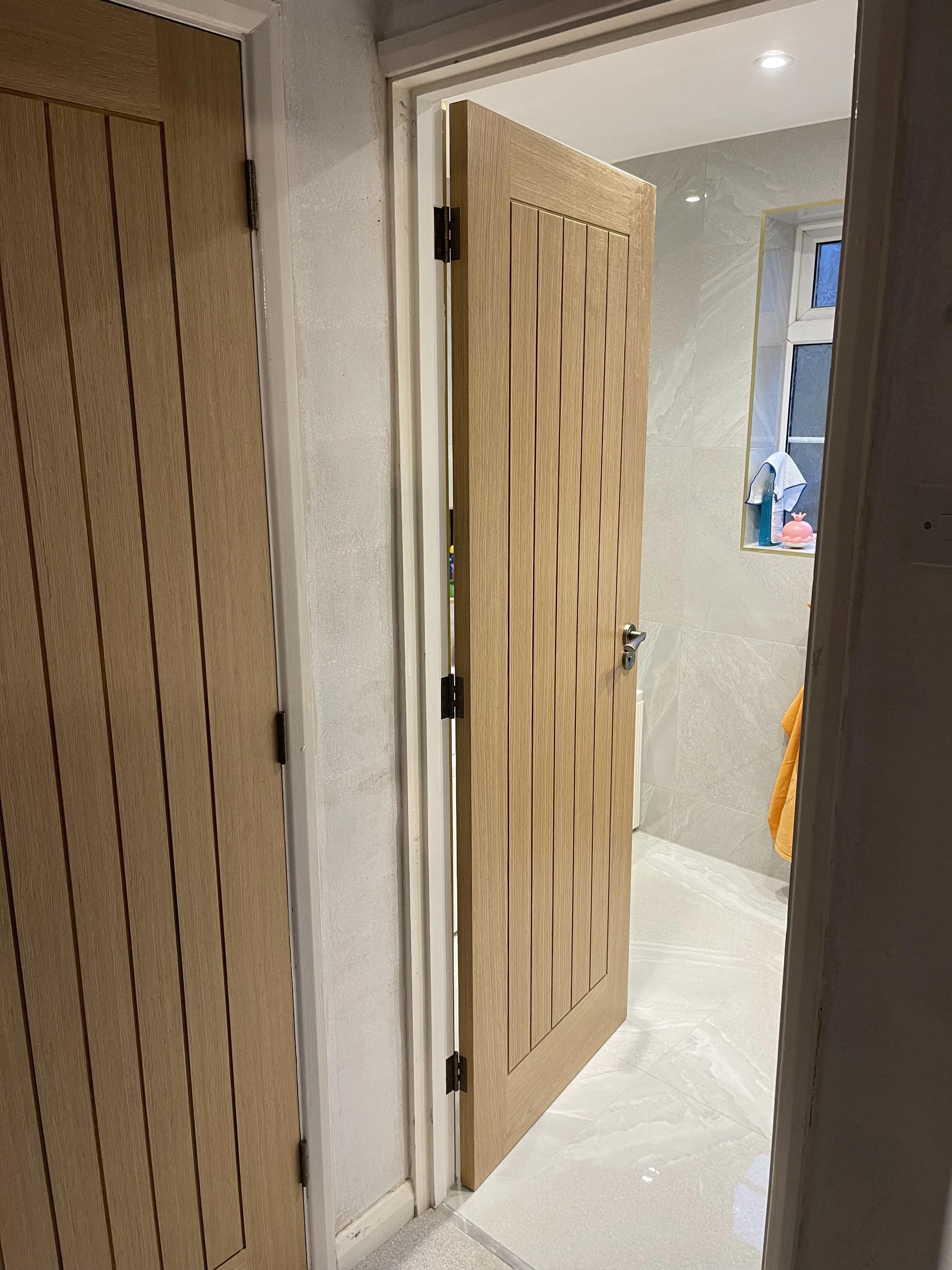 Open bathroom with new oak door revealing tiled walls, a window, and bathroom items like a towel, soap dispenser, and decorated container.