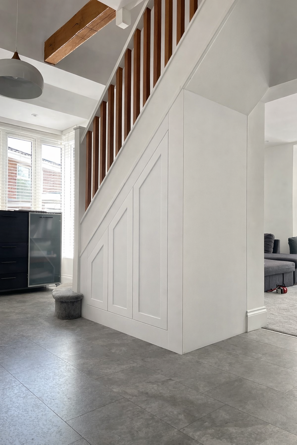 Interior view of a modern home showing a staircase with under stair storage, white paneling and wooden railing, grey tile flooring, a black cabinet near a large window with blinds, and part of a living room with dark grey sofas.