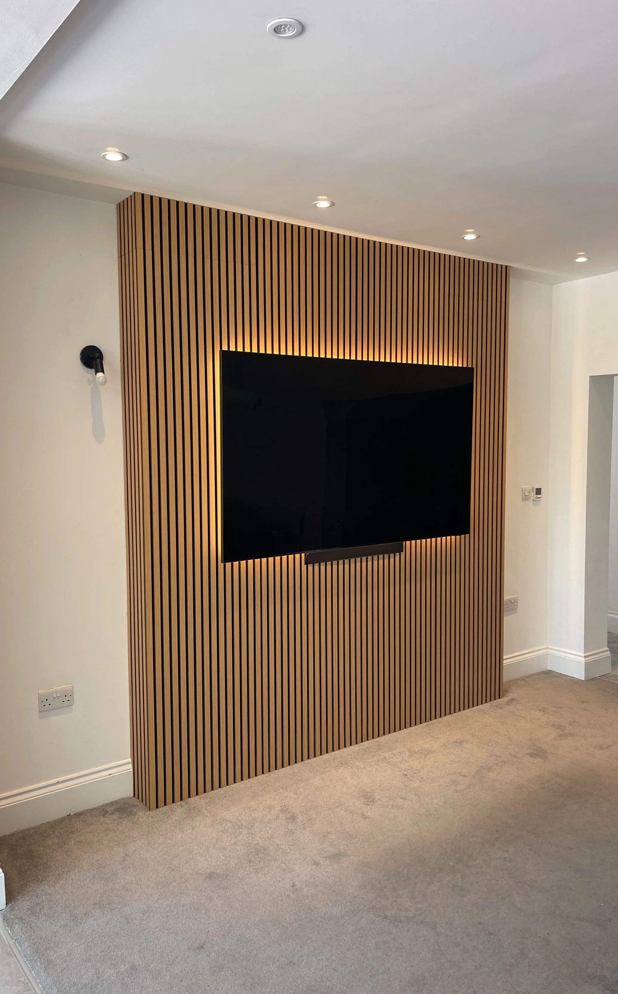 Modern living room with flat-screen TV mounted on a wooden panelled slat media wall, recessed ceiling lights, light-colored carpet, and white walls.