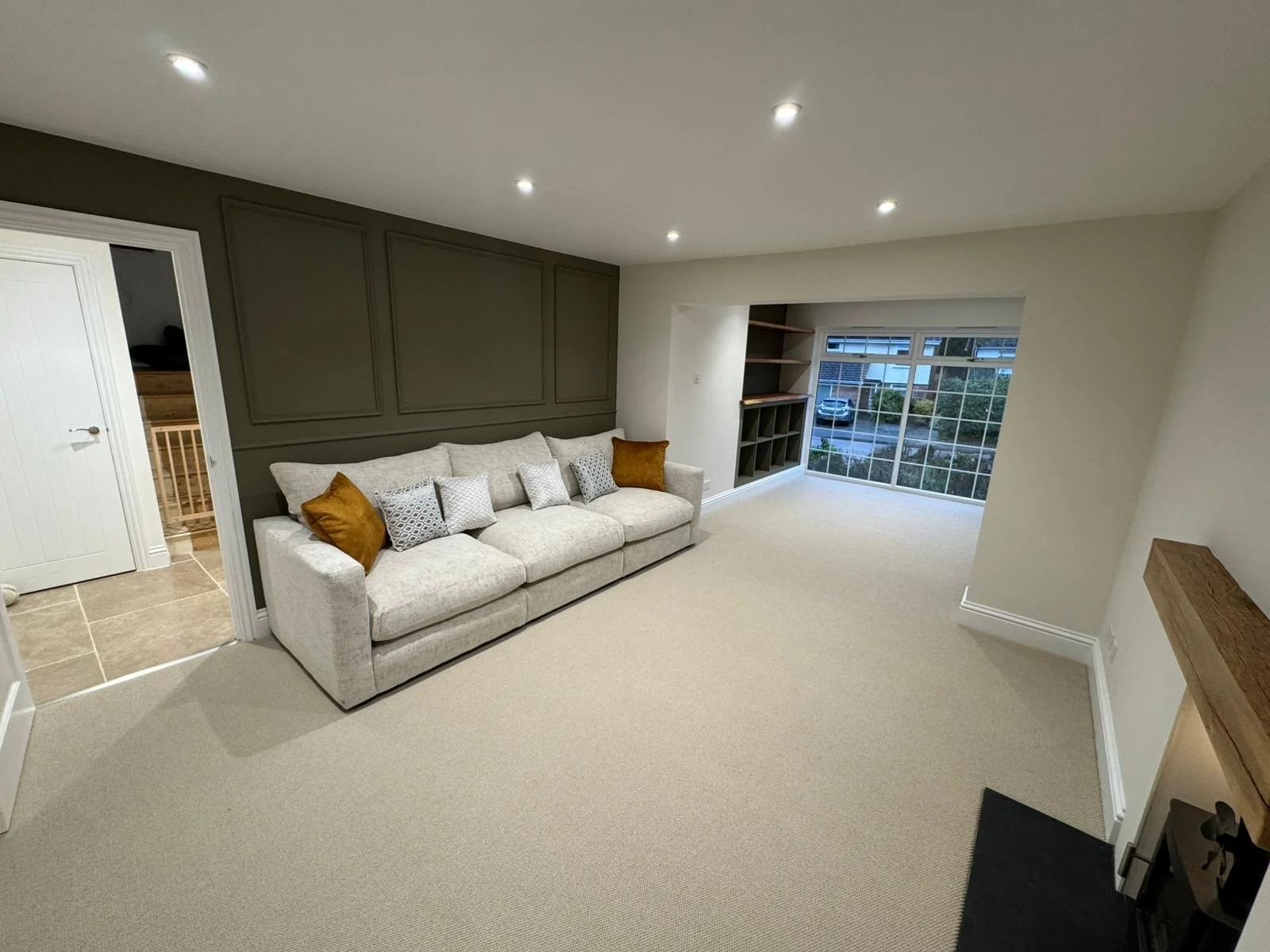 Living room with beige carpet, a large beige sofa with multiple pillows, a dark green accent panel wall behind the sofa, oak custom made shelving and a large window with a view of parked cars outside.