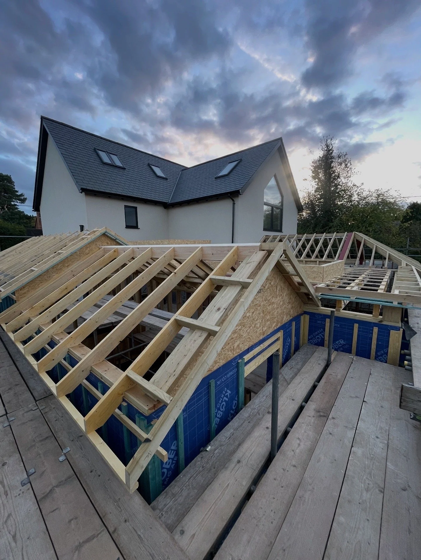 Construction site of a house with wooden framework on a roof, in front of a finished modern white house under a cloudy sky.